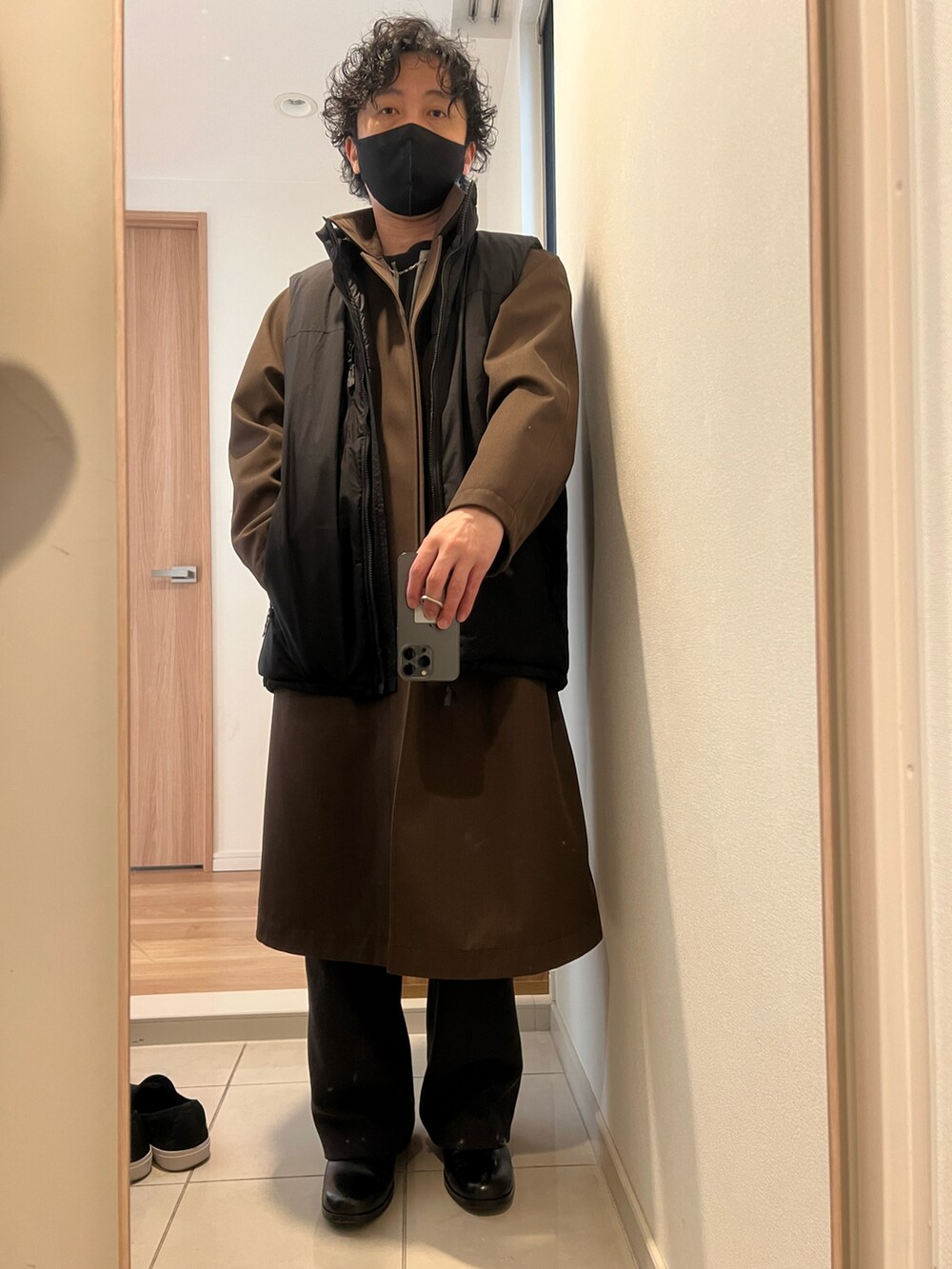ブラウン系のステンカラーコート、ブラック系のダウンベスト、ブラック系のニット/セーターを着用したメンズの冬コーディネートの2枚目の写真