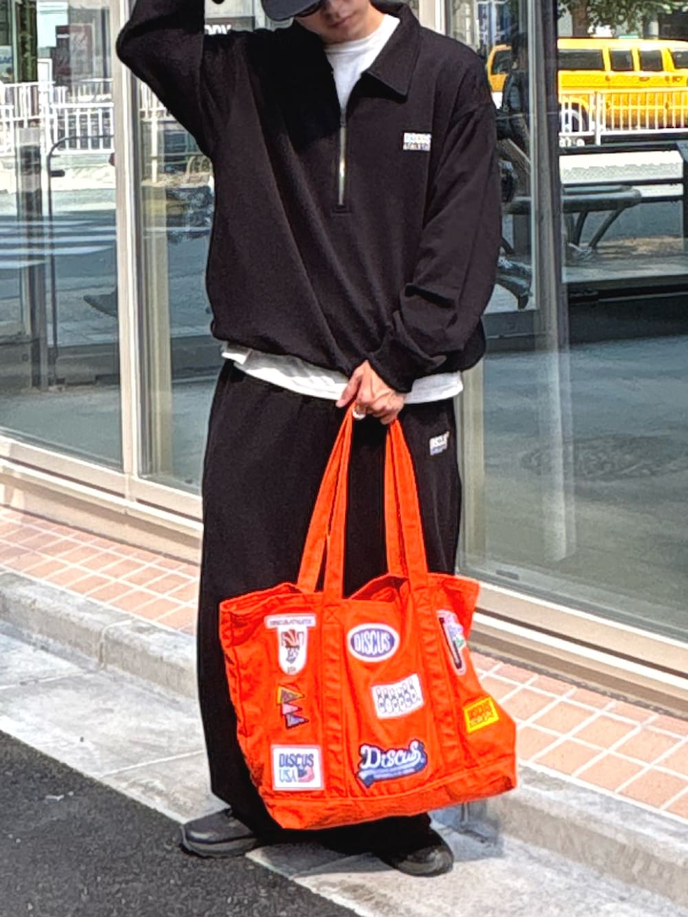 ブラック系のスウェット、ホワイト系のTシャツ/カットソー、オレンジ系のトートバッグを着用したメンズの夏コーディネートの2枚目の写真