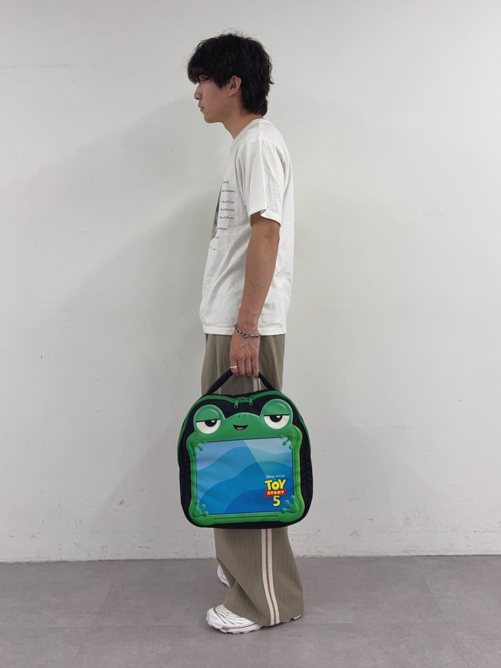 ホワイト系のTシャツ/カットソー、ベージュ系のスラックス、ブラック系のPC・タブレットケースを着用したメンズの春コーディネートの2枚目の写真