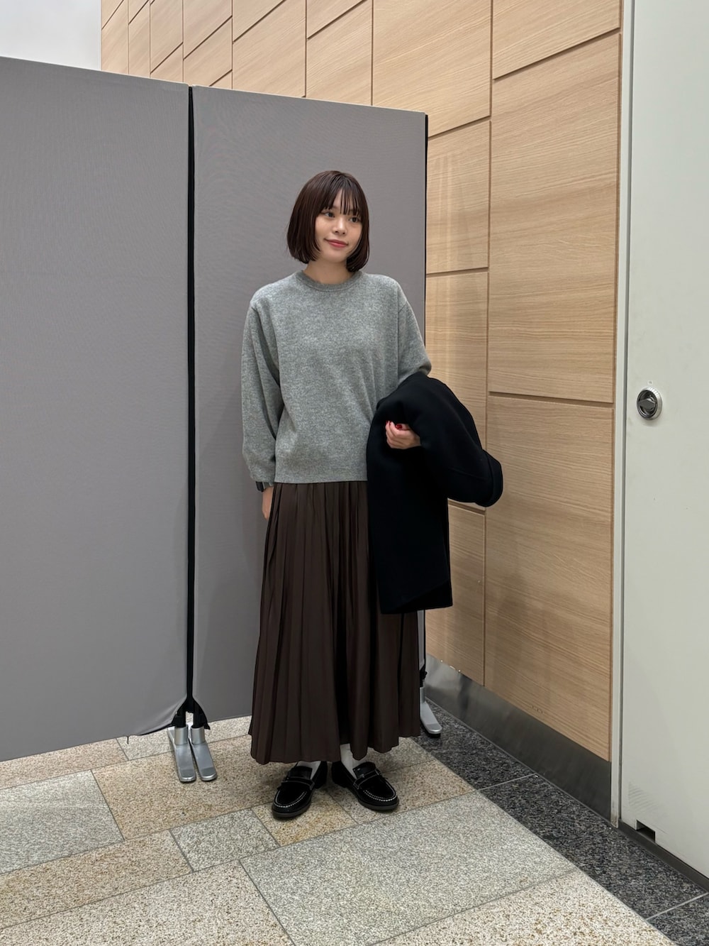 ブラック系のステンカラーコート、ブラウン系のスカート、グレー系のニット/セーターを着用したレディースの秋コーディネートの4枚目の写真