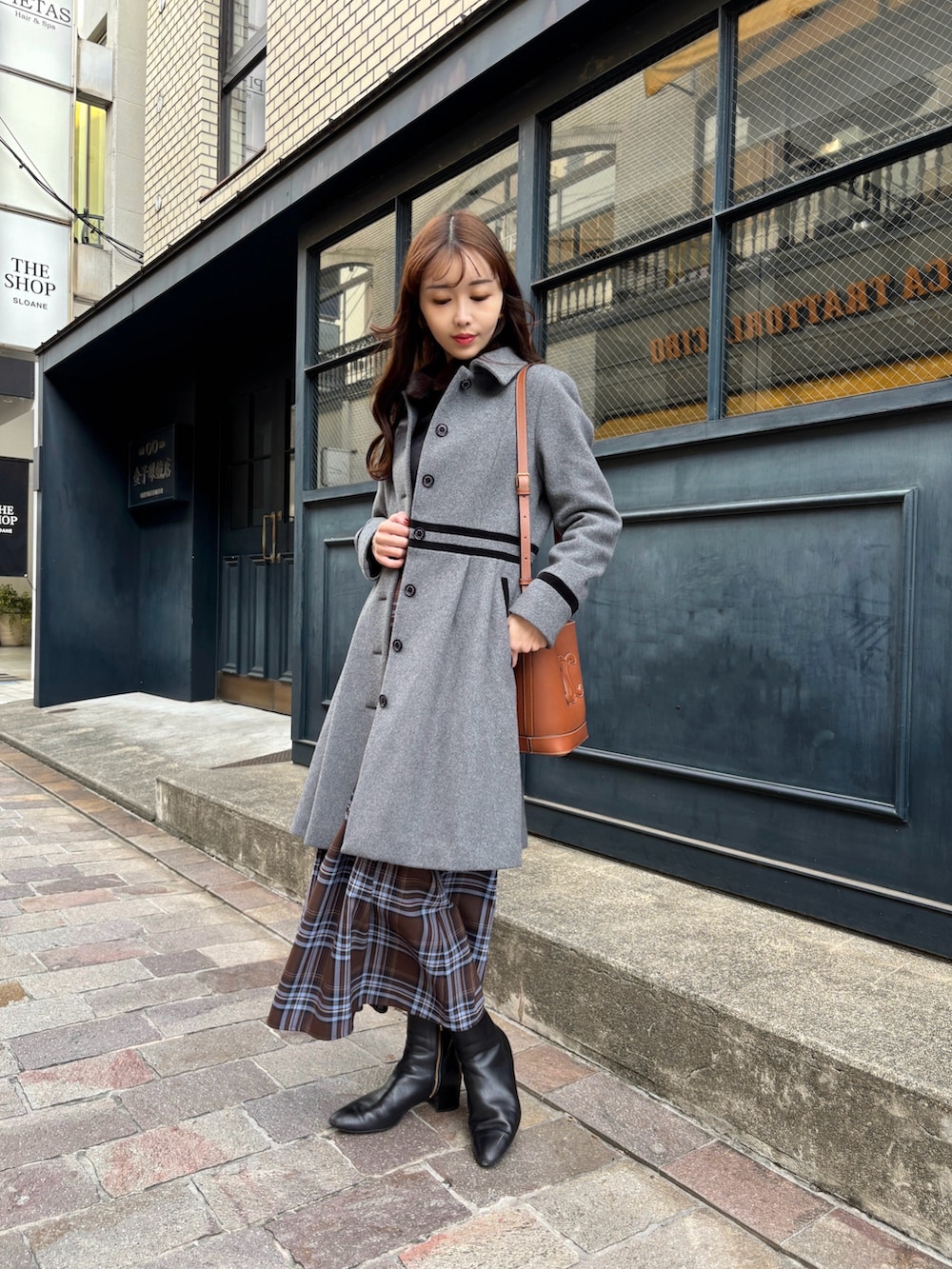 グレー系のステンカラーコート、ブラウン系のスカート、ブラック系のブーツを着用したレディースの冬コーディネートの2枚目の写真