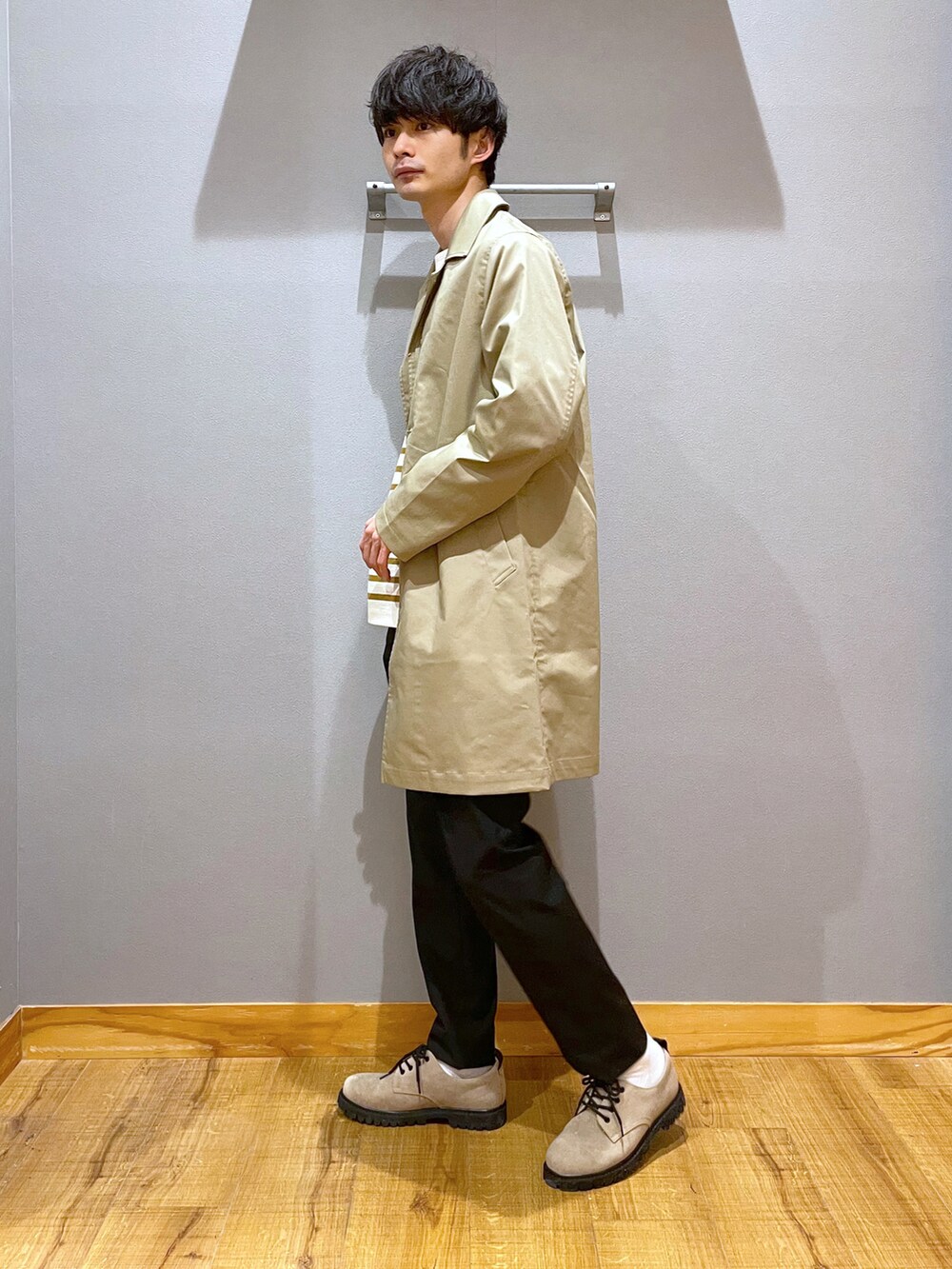 ブラック系のスラックス、ベージュ系のステンカラーコート、ブラウン系のTシャツ/カットソーを着用したメンズの春コーディネートの2枚目の写真