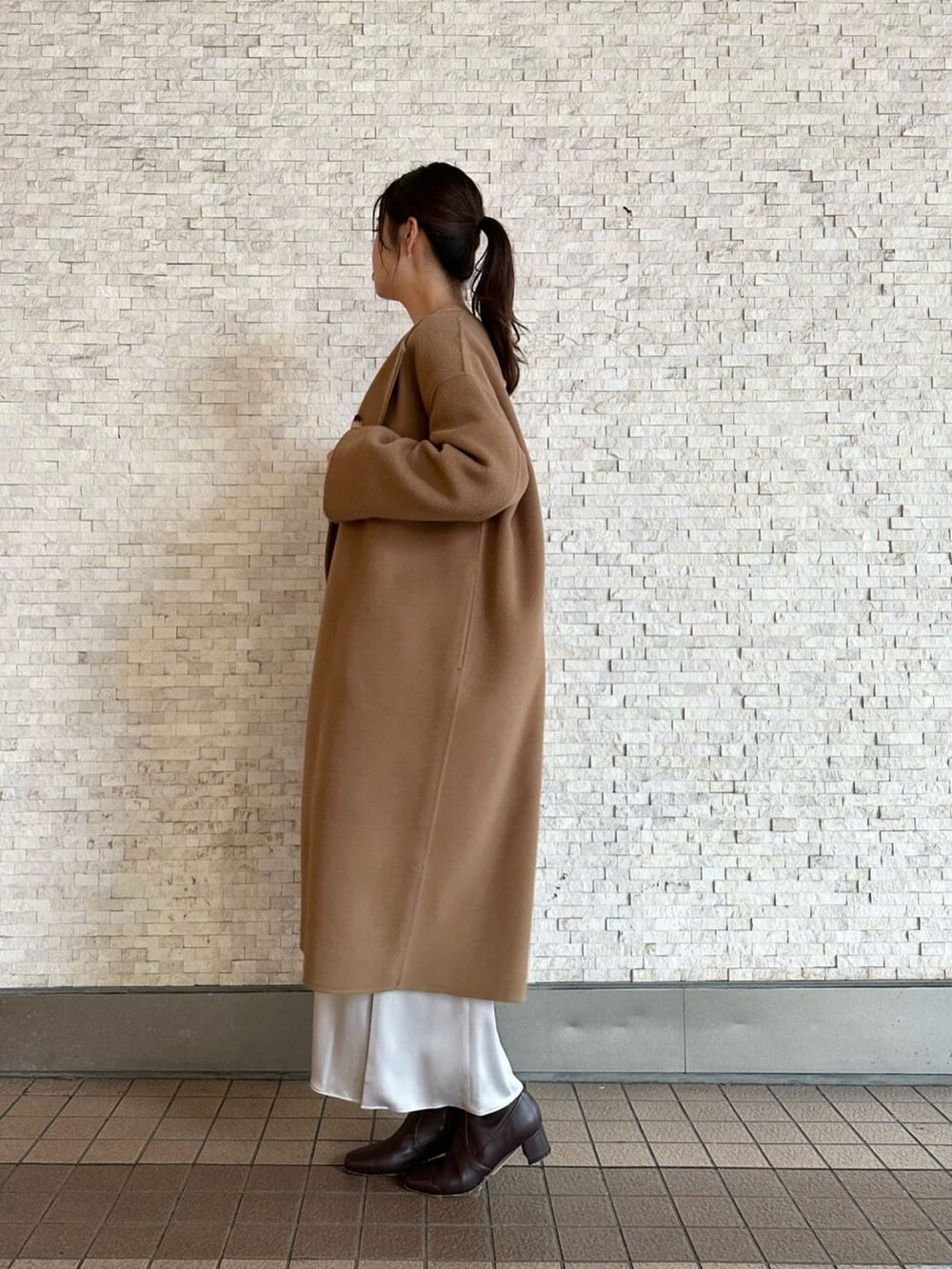 イエロー系のノーカラーコート、グレー系のニット/セーター、ブラウン系のブーツを着用したレディースの秋コーディネートの2枚目の写真