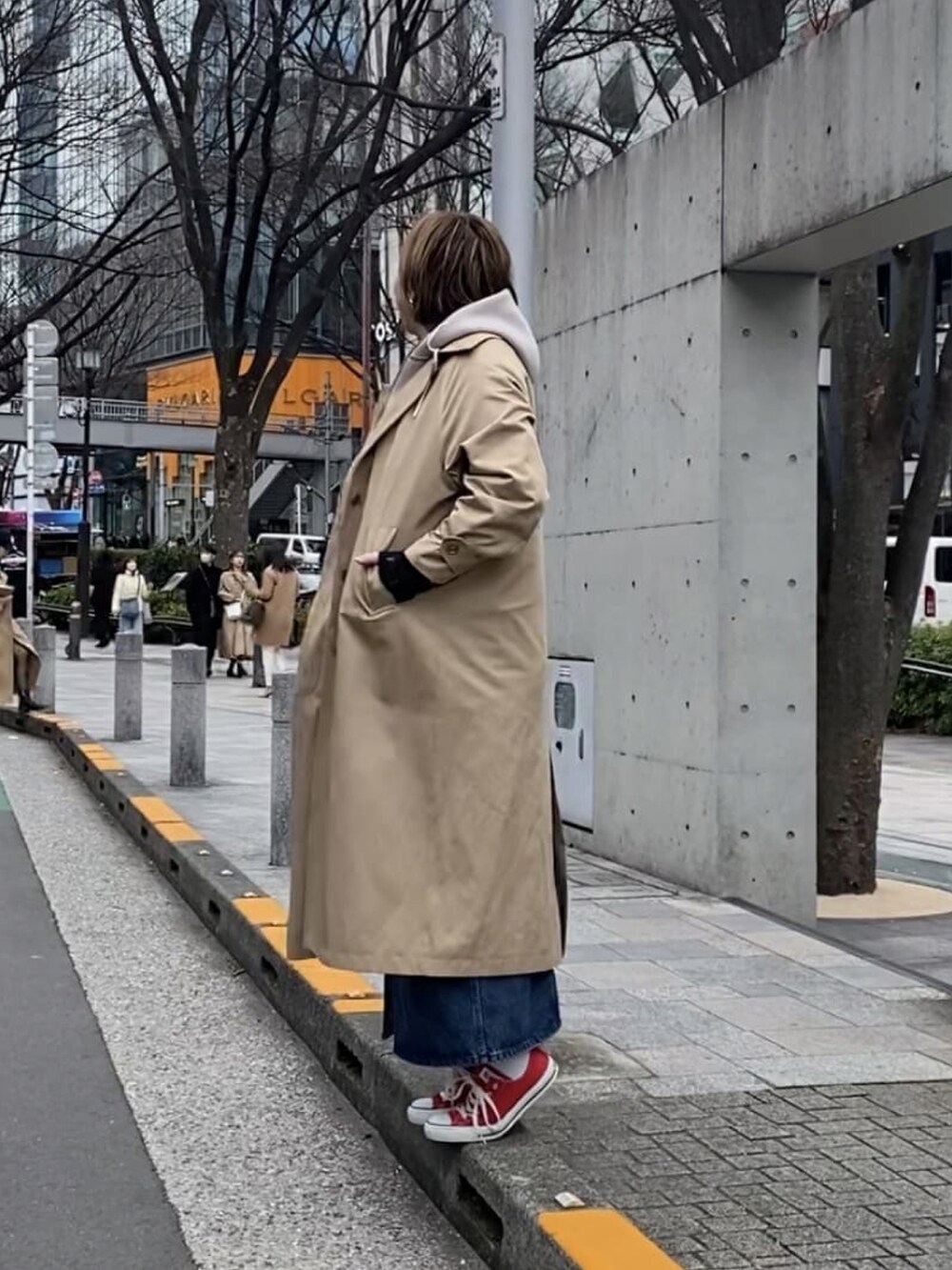 ベージュ系のステンカラーコート、ブルー系のジャンパースカート、ブラック系のTシャツ/カットソーを着用したレディースの冬コーディネートの2枚目の写真