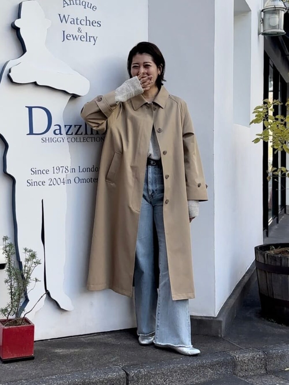 ベージュ系のステンカラーコート、ブルー系のデニムパンツ、グレー系のニット/セーターを着用したレディースの冬コーディネートの2枚目の写真