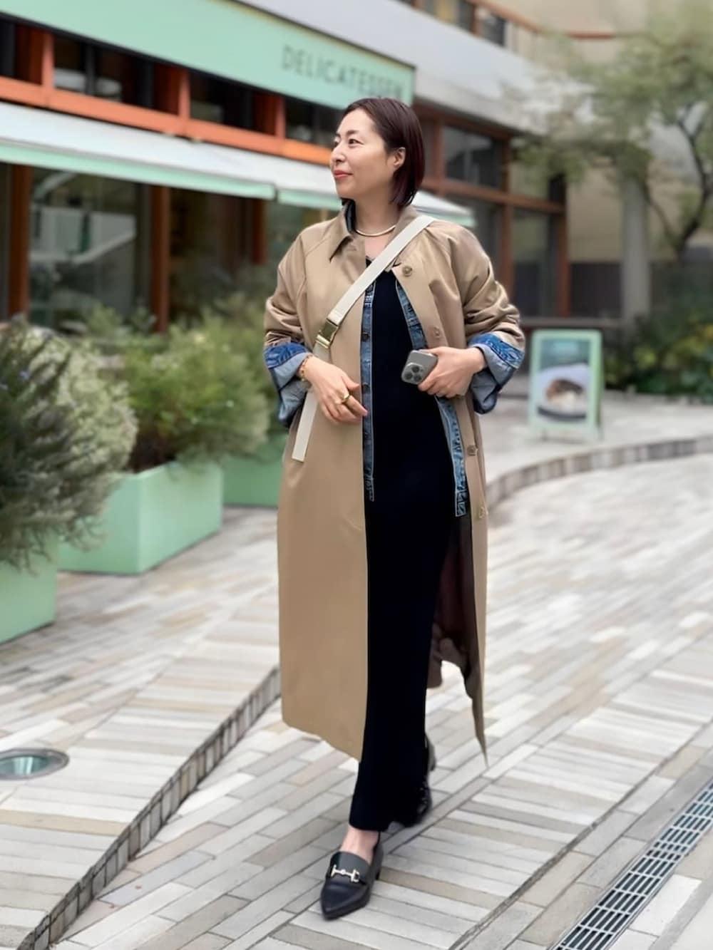 ベージュ系のステンカラーコート、ホワイト系のボディバッグ/ウエストポーチ、ブラック系のワンピースを着用したレディースの冬コーディネートの2枚目の写真