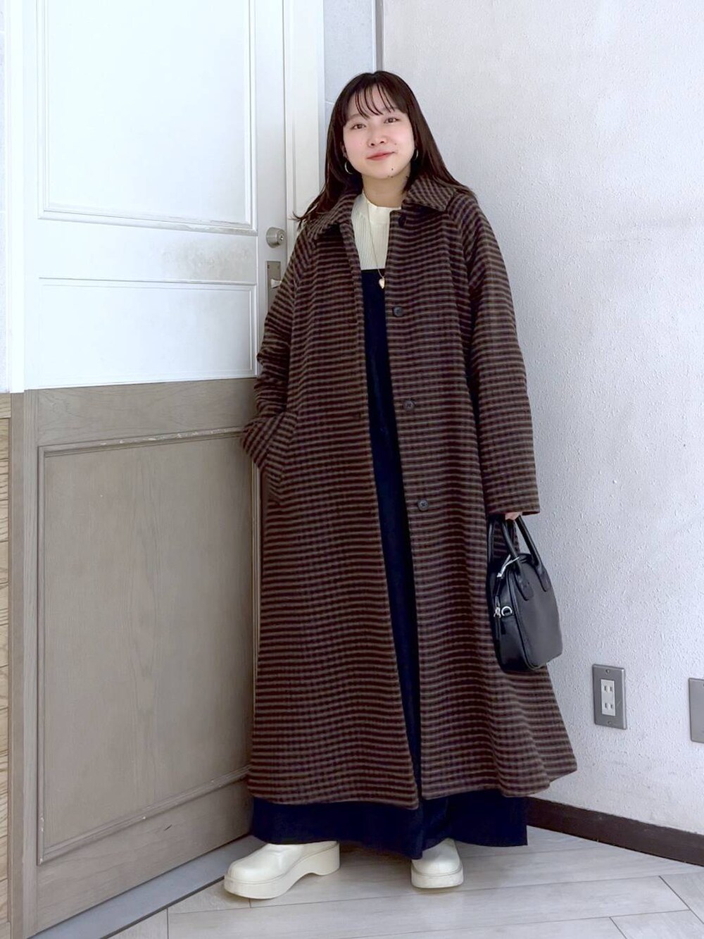 ブラウン系のノーカラーコート、ホワイト系のブーツ、ブラック系のワンピースを着用したレディースの冬コーディネートの2枚目の写真