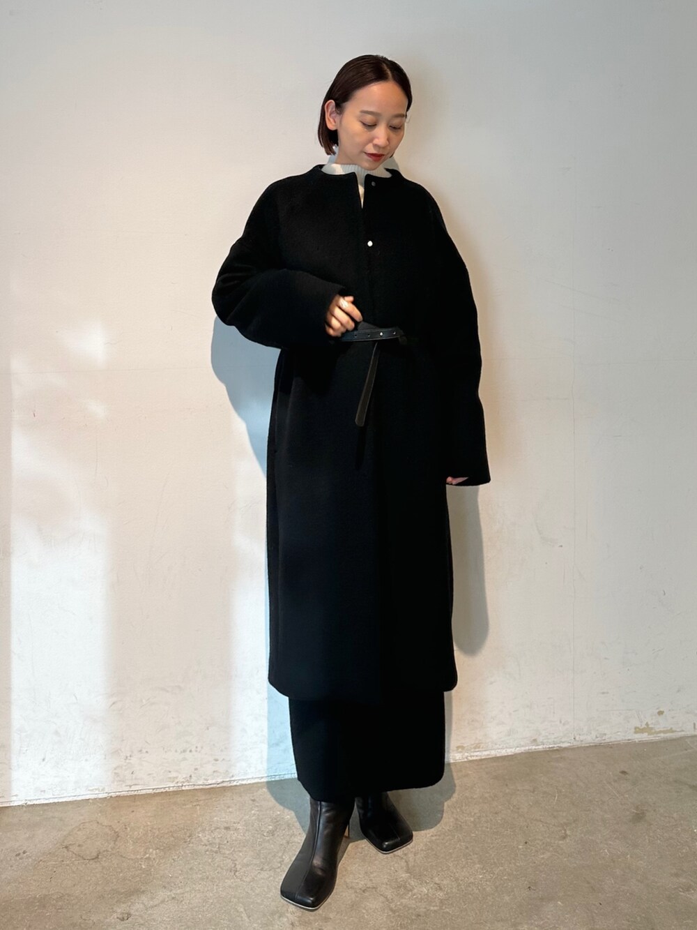 ブラック系のノーカラーコート、ブラック系のブーツ、ホワイト系のニット/セーターを着用したレディースの秋コーディネートの2枚目の写真