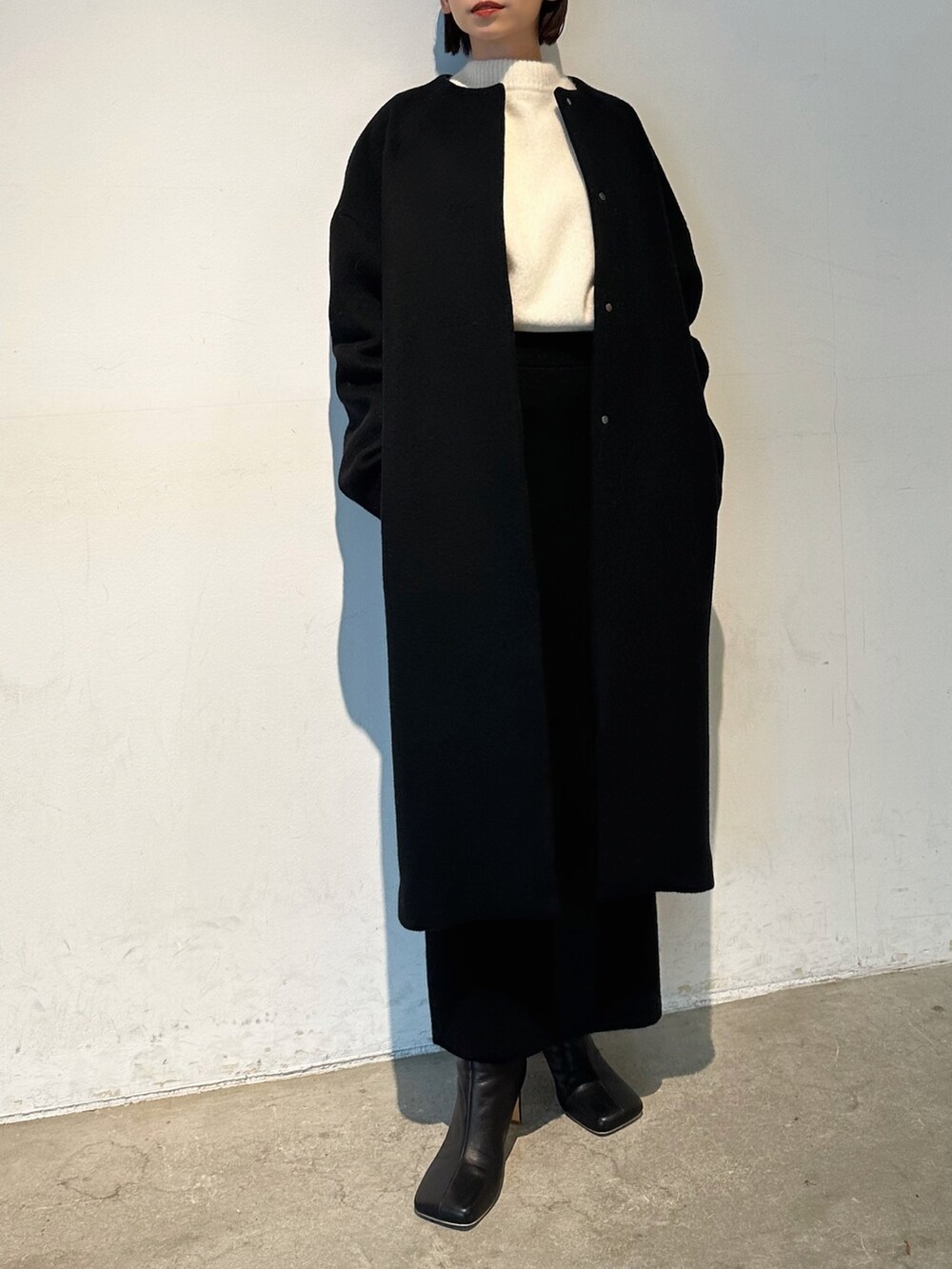 ブラック系のノーカラーコート、ブラック系のブーツ、ホワイト系のニット/セーターを着用したレディースの秋コーディネートの3枚目の写真