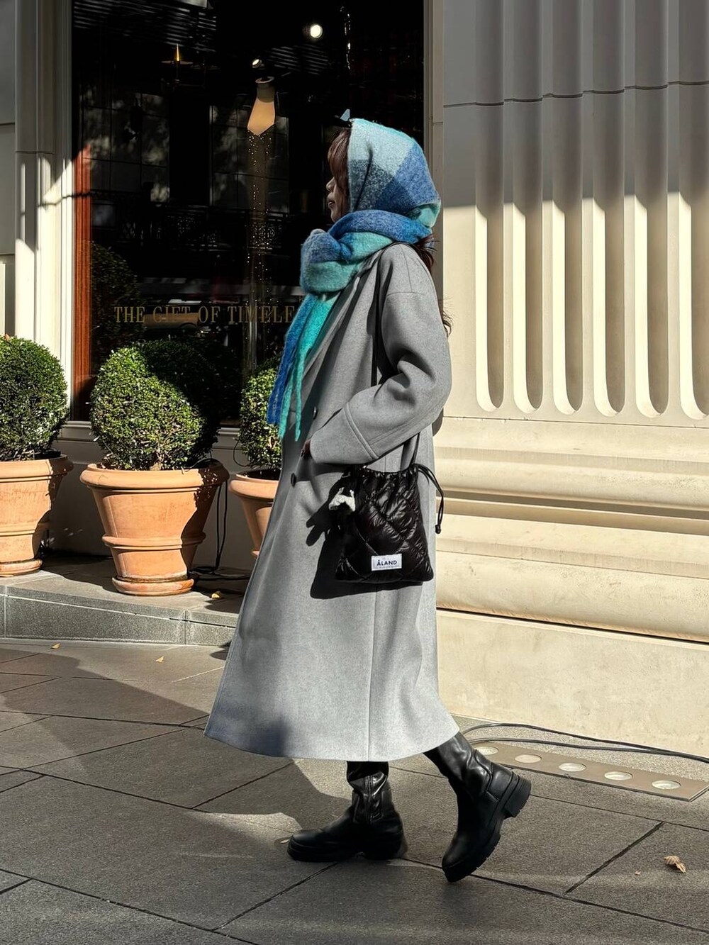 グレー系のチェスターコート、ブラック系のショルダーバッグ、ブルー系のマフラーを着用したレディースの冬コーディネートの2枚目の写真