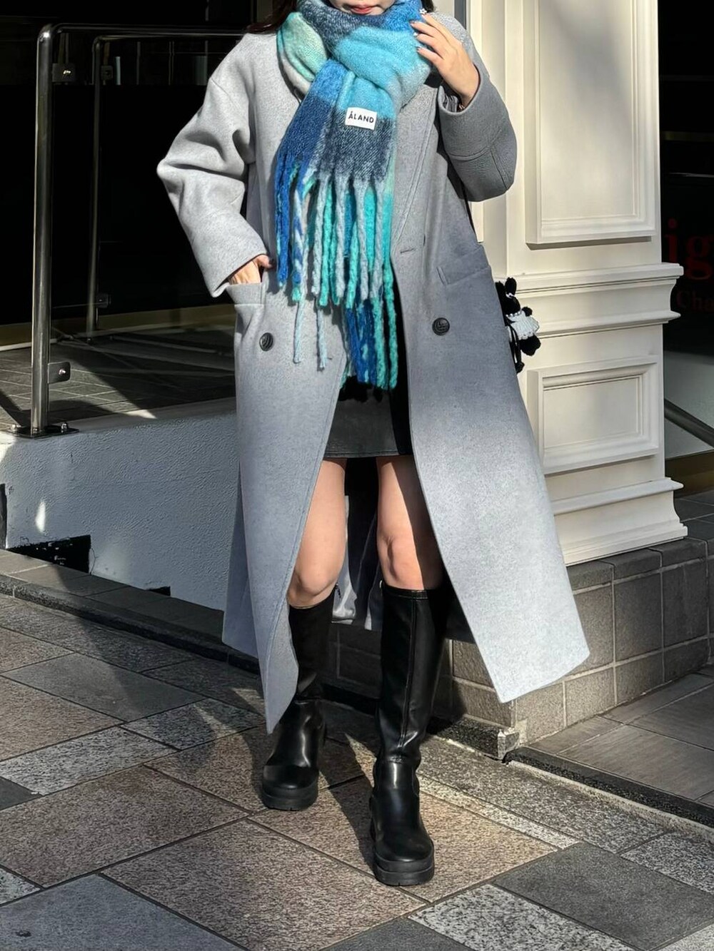 グレー系のチェスターコート、ブラック系のショルダーバッグ、ブルー系のマフラーを着用したレディースの冬コーディネートの5枚目の写真