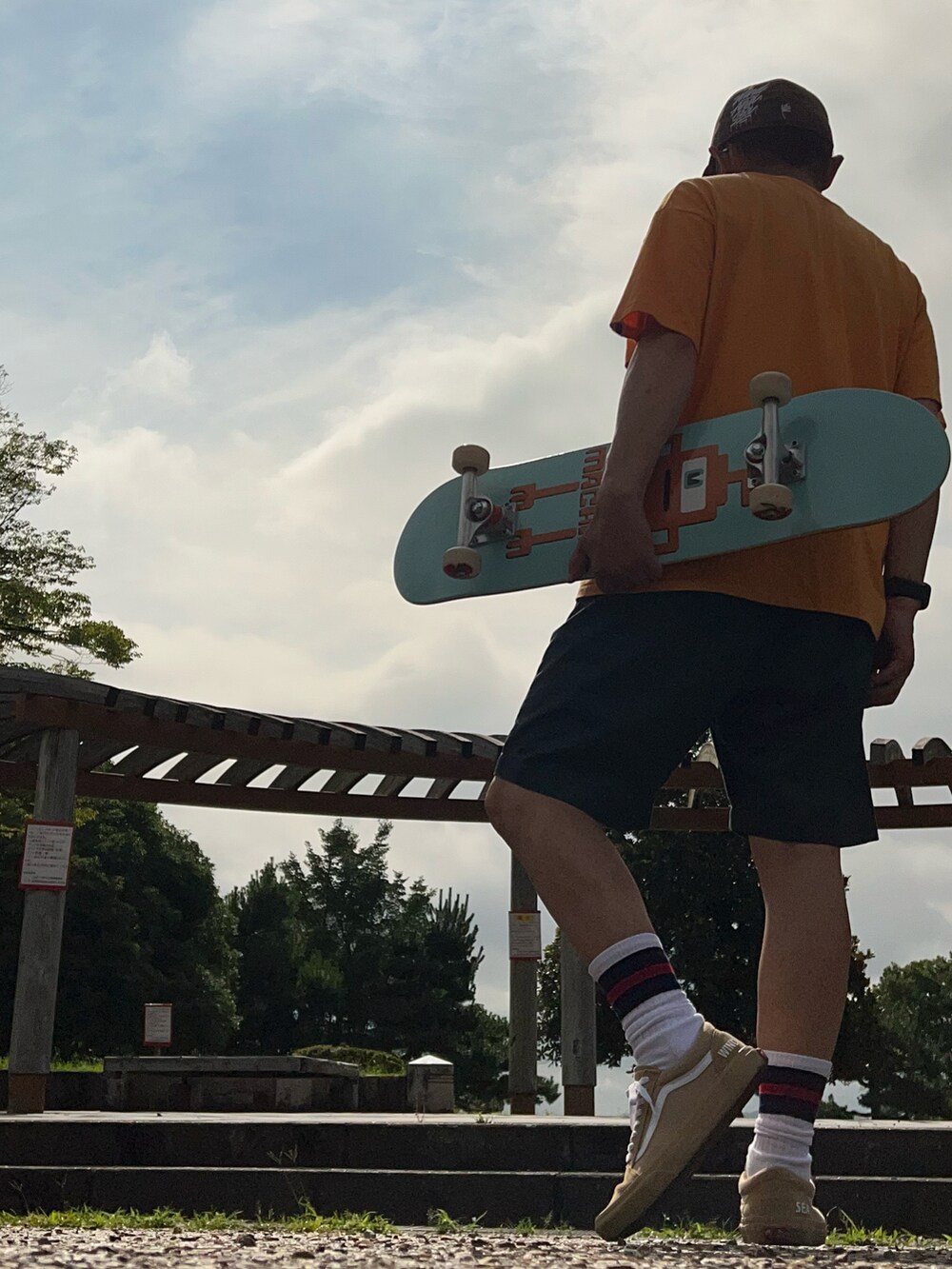 オレンジ系のTシャツ/カットソー、グリーン系のチノパンツ、ベージュ系のスニーカーを着用したメンズの夏コーディネートの2枚目の写真