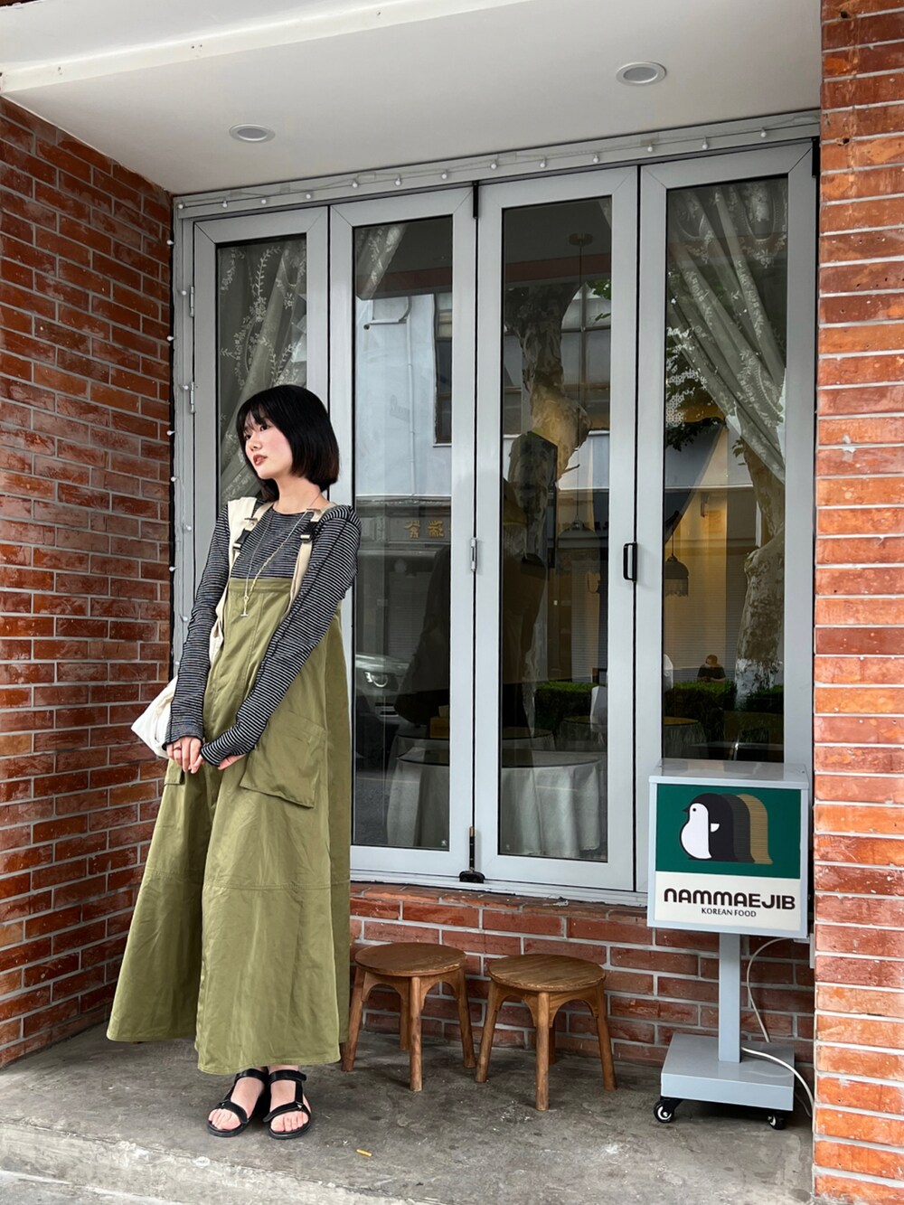 グリーン系のジャンパースカート、ベージュ系のトートバッグ、ブラック系のTシャツ/カットソーを着用したレディースの夏コーディネートの2枚目の写真