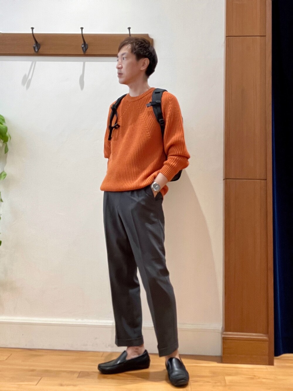 グレー系のスラックス、ブラック系のバックパック/リュック、オレンジ系のニット/セーターを着用したメンズの夏コーディネートの3枚目の写真