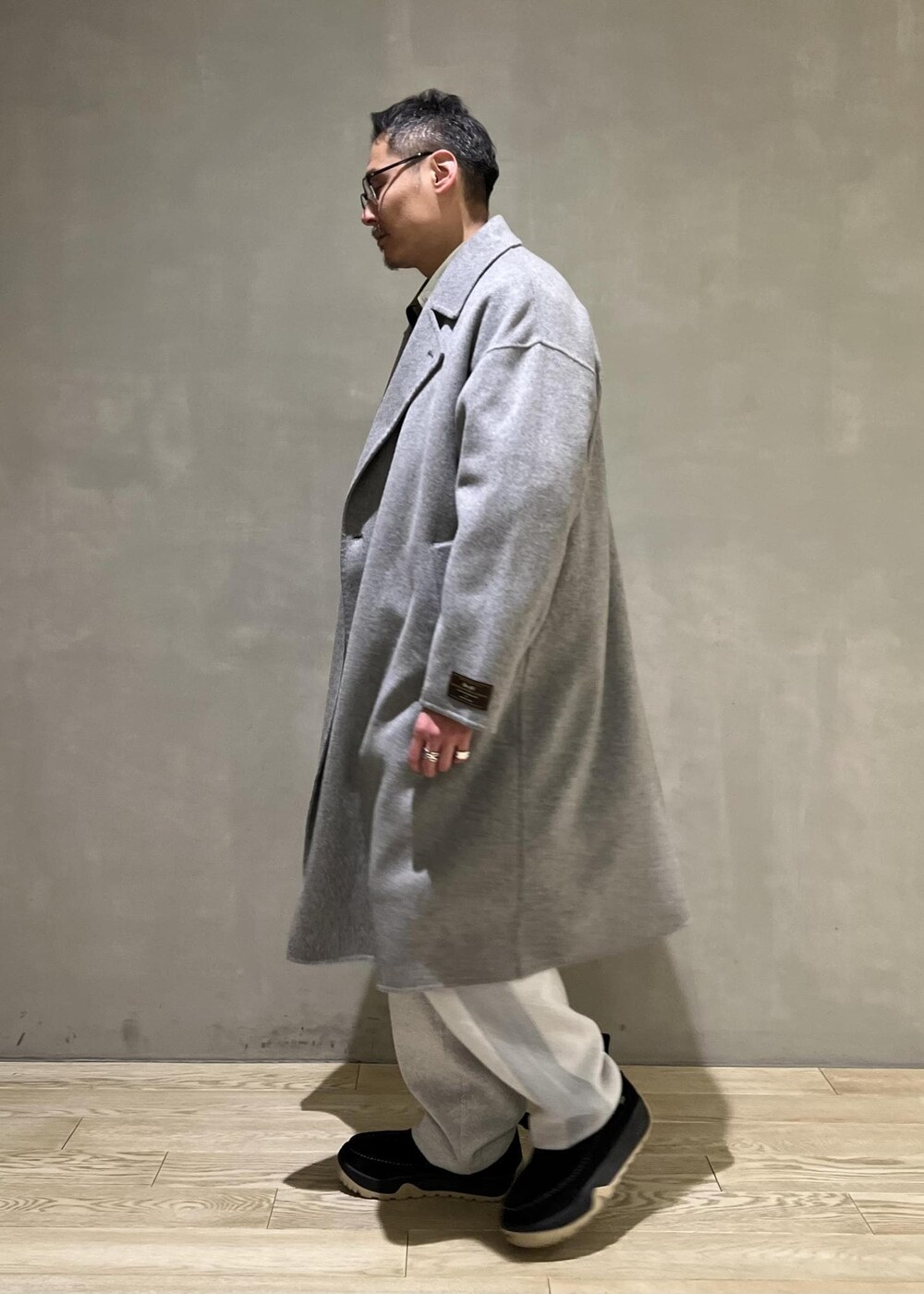 グレー系のステンカラーコート、グレー系のその他パンツ、ブラック系のショルダーバッグを着用したメンズの冬コーディネートの2枚目の写真