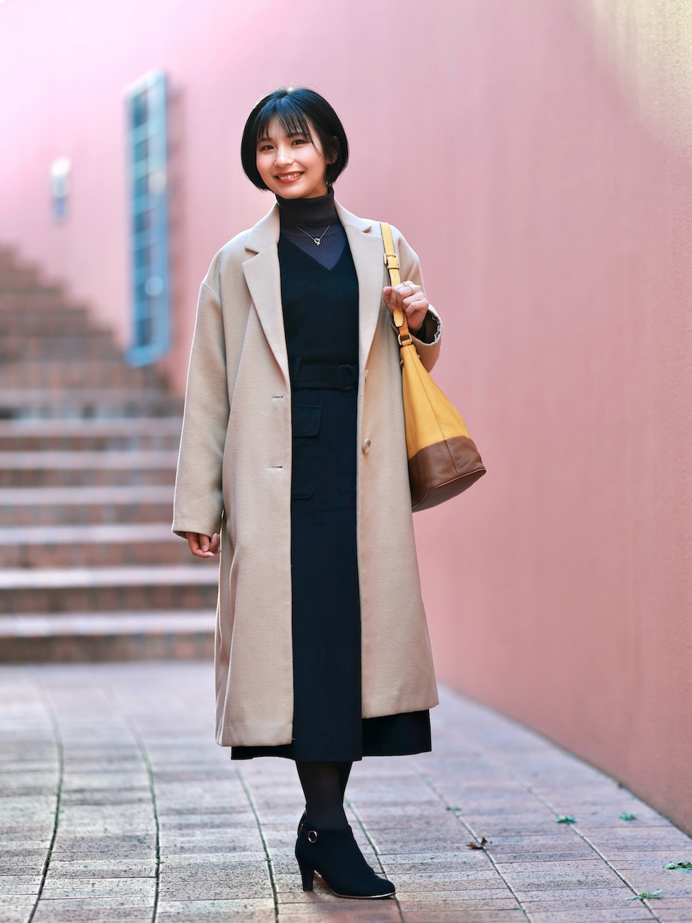 ベージュ系のチェスターコート、ブラック系のジャンパースカート、ブラック系のブーティを着用したレディースの冬コーディネートの2枚目の写真