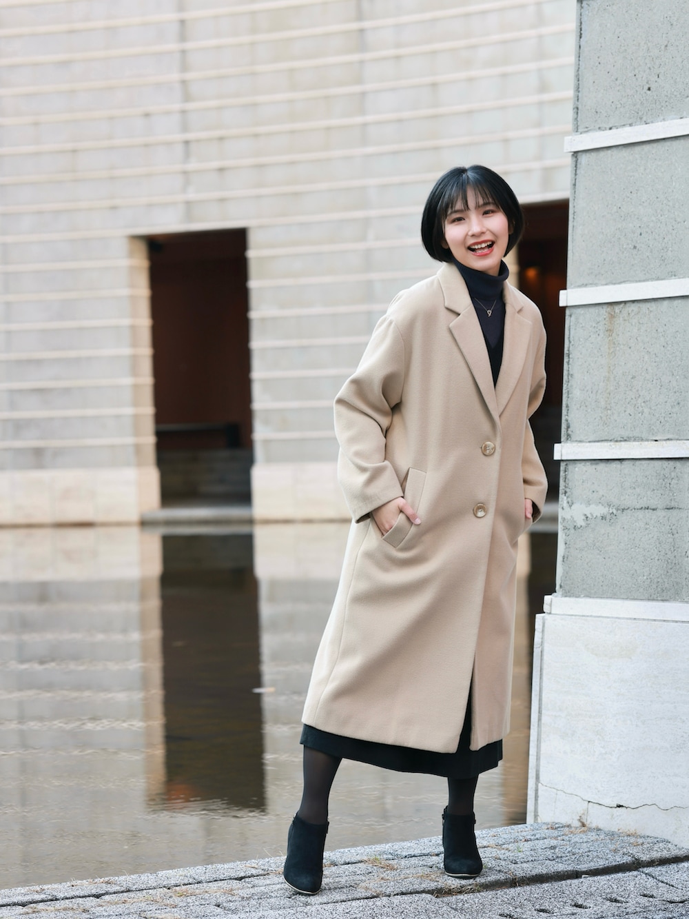 ベージュ系のチェスターコート、ブラック系のジャンパースカート、ブラック系のブーティを着用したレディースの冬コーディネートの3枚目の写真