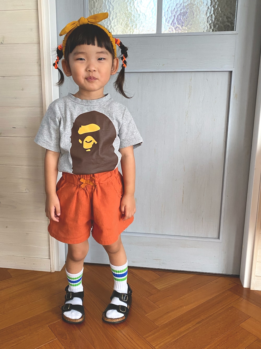 オレンジ系のパンツ、ブラック系のサンダル、イエロー系のヘアバンドを着用したキッズの夏コーディネートの2枚目の写真