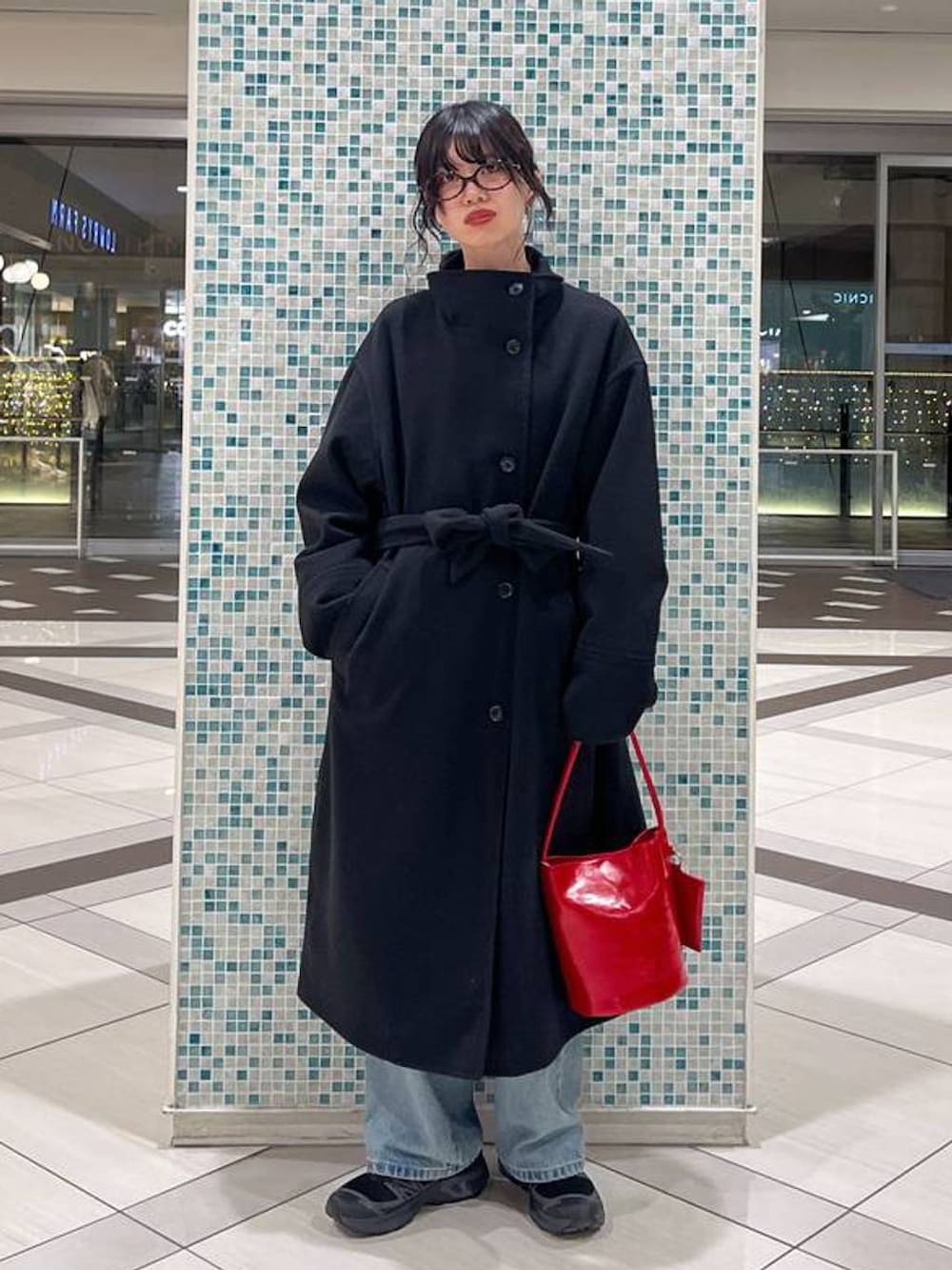 ブラック系のステンカラーコート、レッド系のトートバッグ、ブルー系のデニムパンツを着用したレディースの冬コーディネートの3枚目の写真