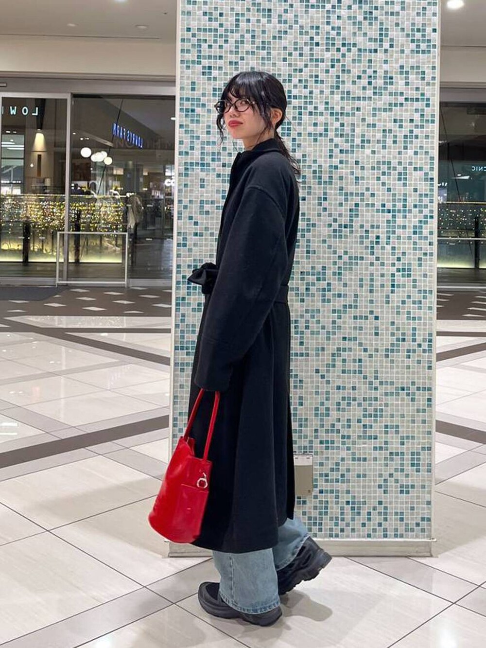 ブラック系のステンカラーコート、レッド系のトートバッグ、ブルー系のデニムパンツを着用したレディースの冬コーディネートの2枚目の写真