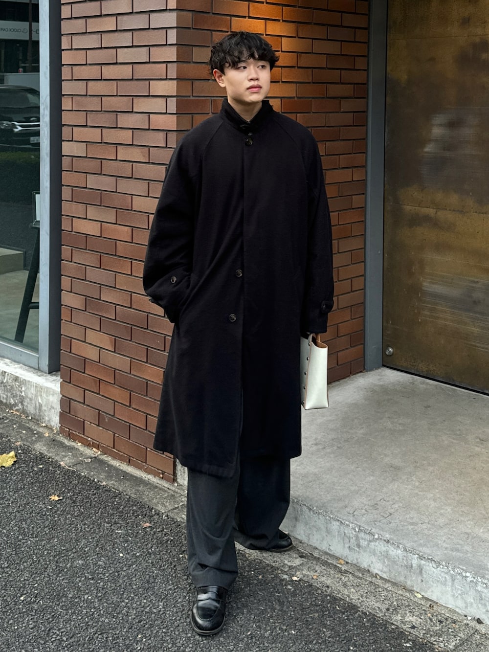 ブラック系のステンカラーコート、ホワイト系のハンドバッグ、グレー系のスラックスを着用したメンズの冬コーディネートの2枚目の写真