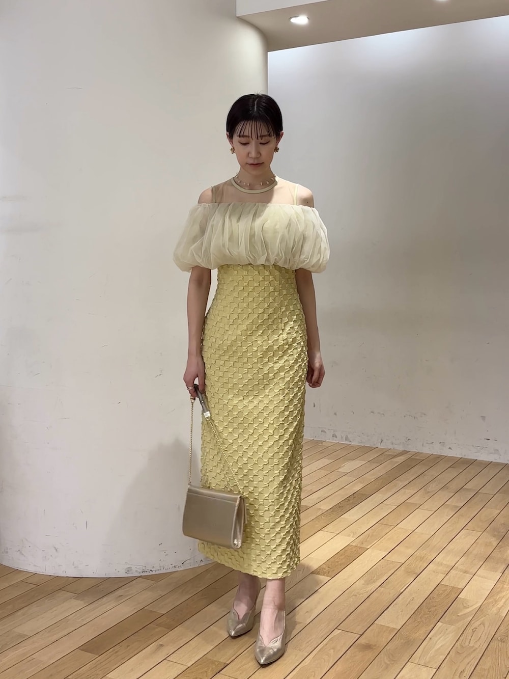 イエロー系のドレス、ゴールド系のパンプス、ゴールド系のクラッチバッグを着用したレディースの春コーディネートの2枚目の写真