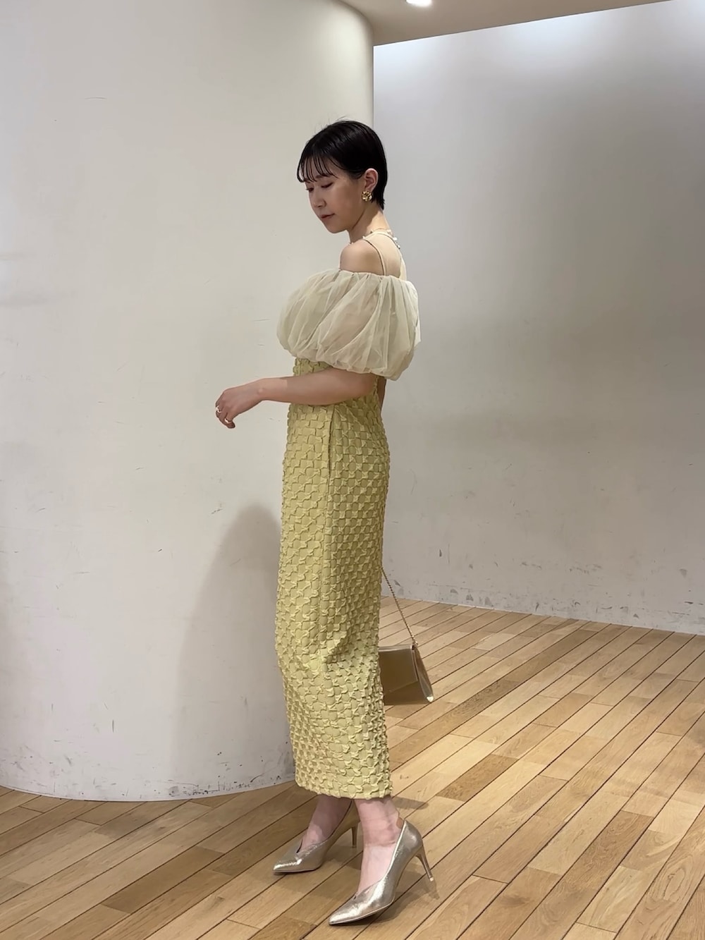 イエロー系のドレス、ゴールド系のパンプス、ゴールド系のクラッチバッグを着用したレディースの春コーディネートの3枚目の写真