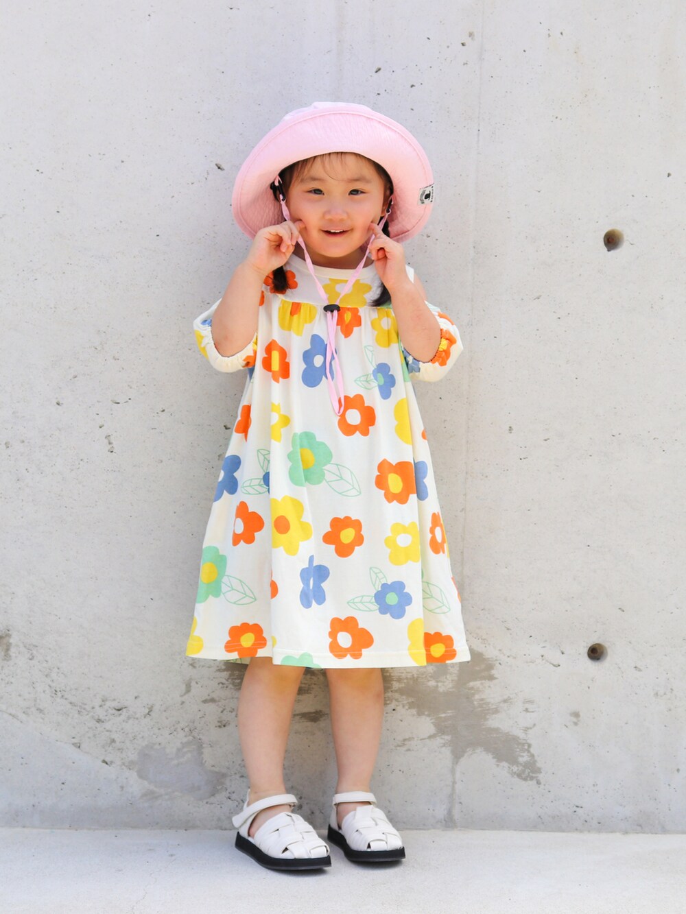 オレンジ系のワンピース、ホワイト系のサンダル、ピンク系のハットを着用したキッズの夏コーディネートの2枚目の写真