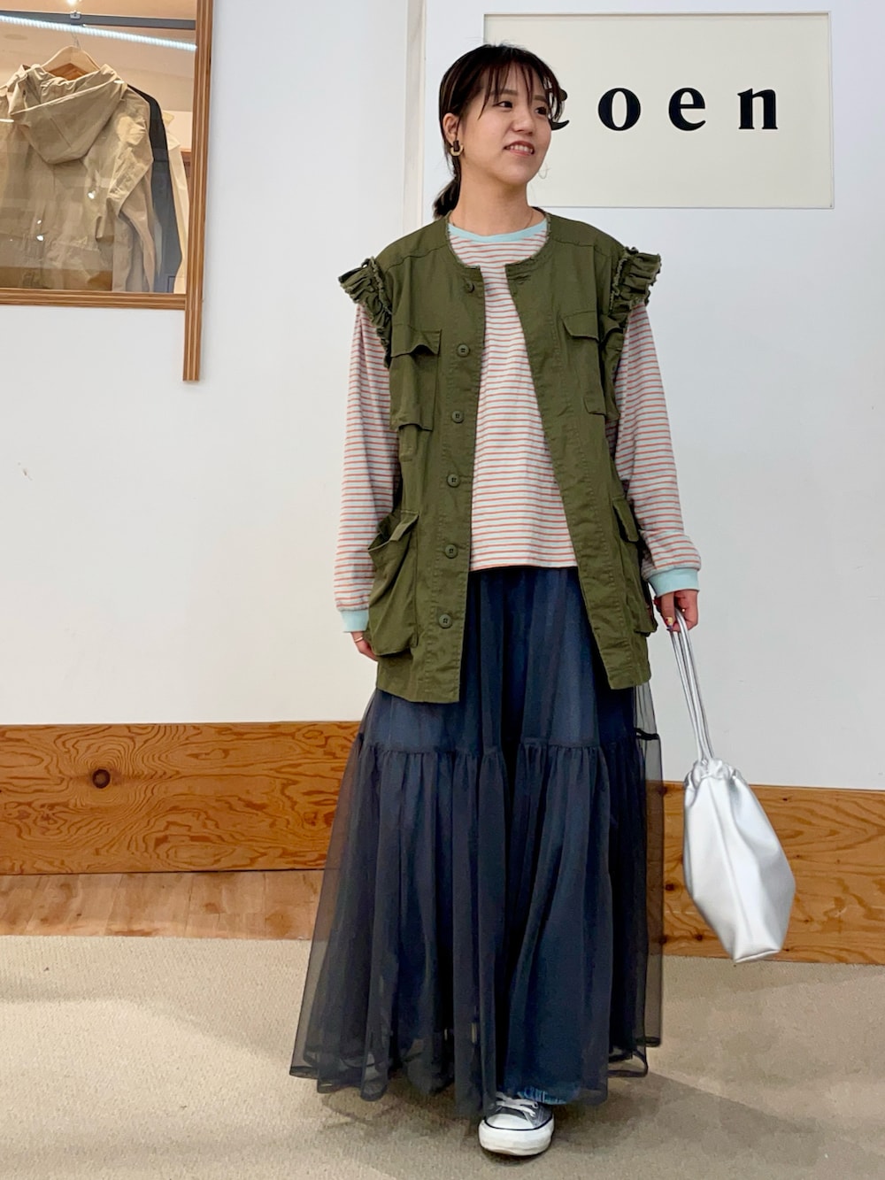 グリーン系のベスト、オレンジ系のTシャツ/カットソー、グレー系のスカートを着用したレディースの春コーディネートの3枚目の写真