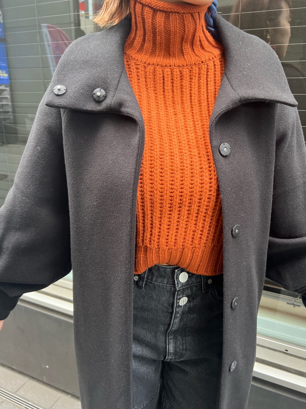 ブラック系のデニムパンツ、オレンジ系のニット/セーター、ブラック系のステンカラーコートを着用したレディースの秋コーディネートの5枚目の写真