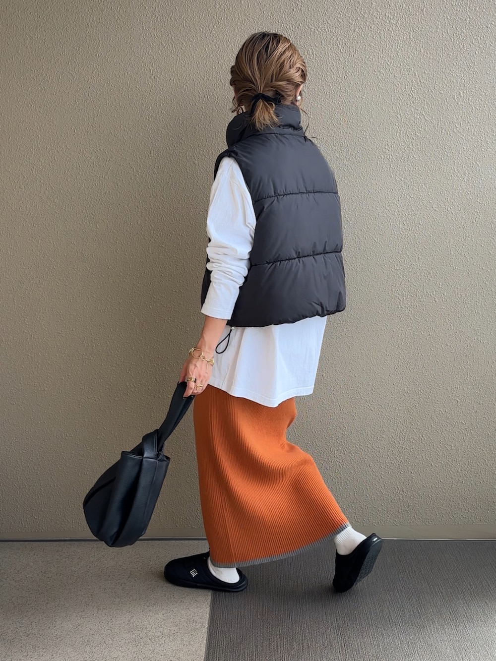 オレンジ系のスカート、ブラック系のダウンベスト、ブラック系のハンドバッグを着用したレディースの秋コーディネートの2枚目の写真