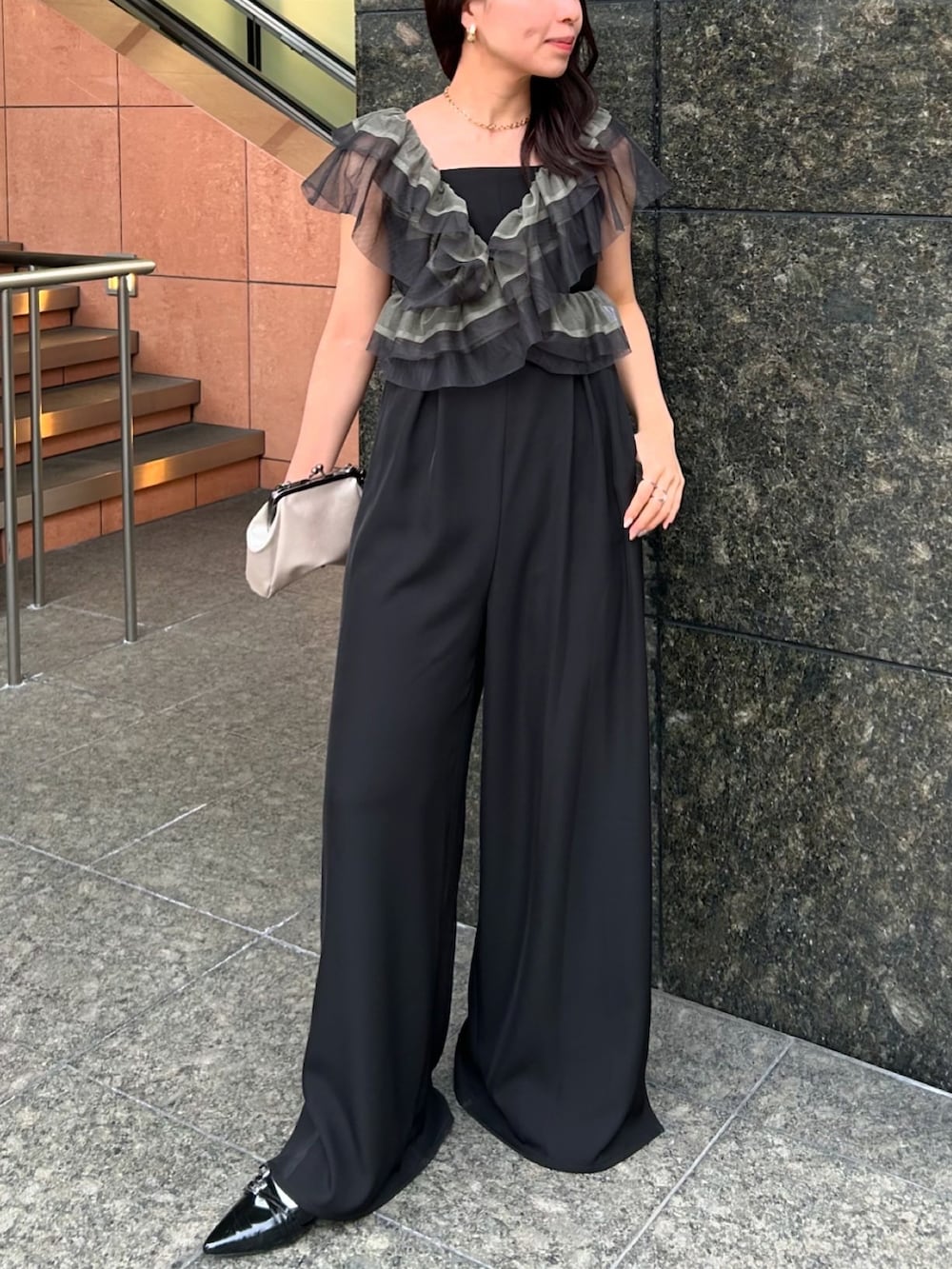 ブラック系のパンツドレス、ゴールド系のネックレス、ゴールド系のピアス（両耳用）を着用したレディースの春コーディネートの2枚目の写真