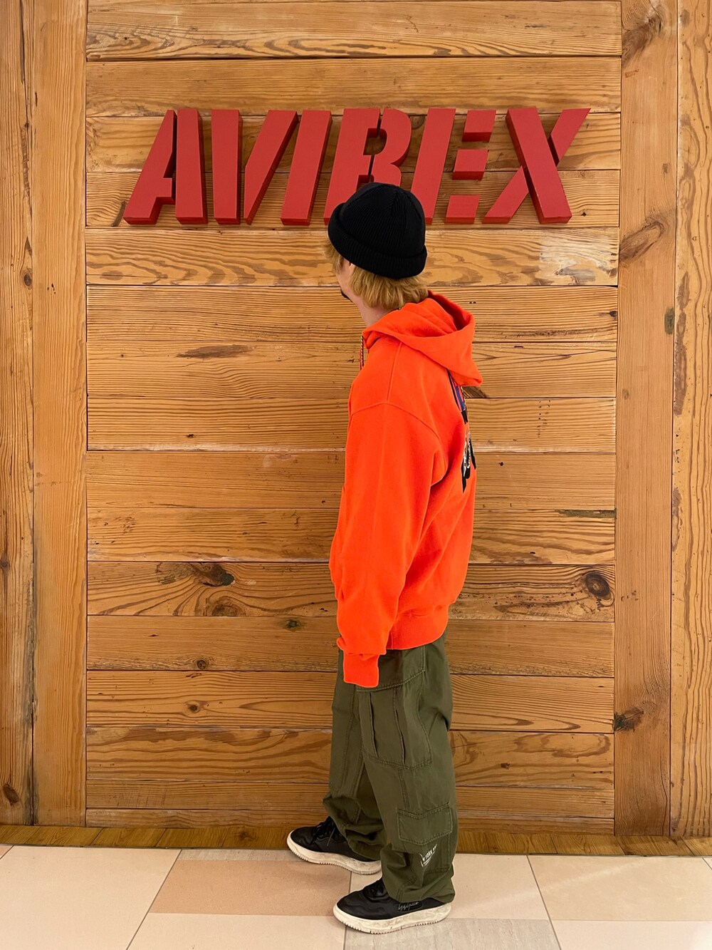 オレンジ系のパーカー、グリーン系のカーゴパンツ、ブラック系のニットキャップ/ビーニーを着用したメンズの夏コーディネートの2枚目の写真