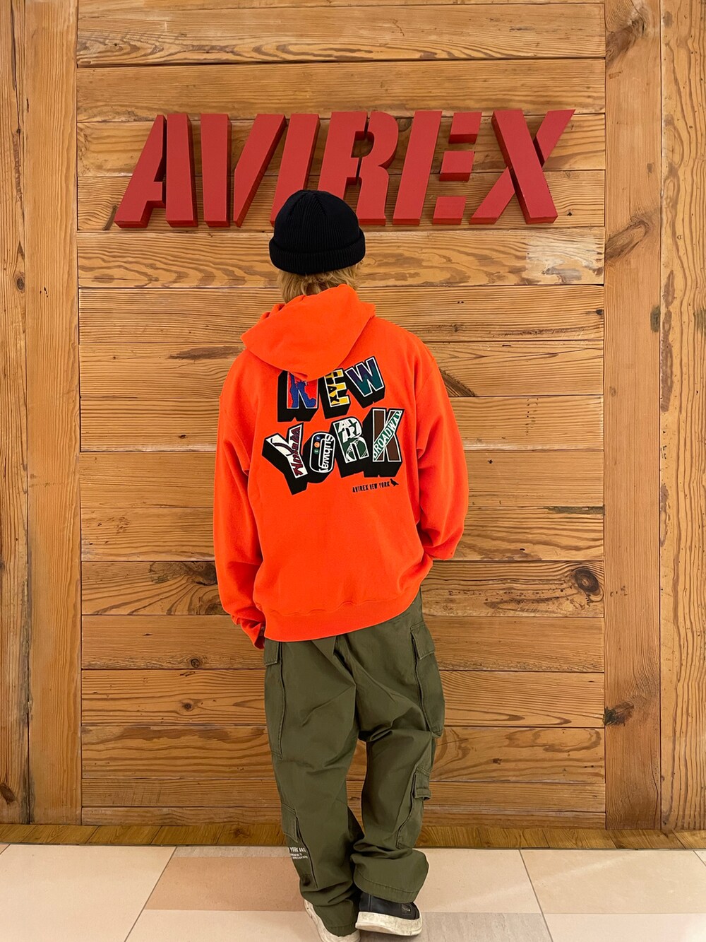 オレンジ系のパーカー、グリーン系のカーゴパンツ、ブラック系のニットキャップ/ビーニーを着用したメンズの夏コーディネートの3枚目の写真