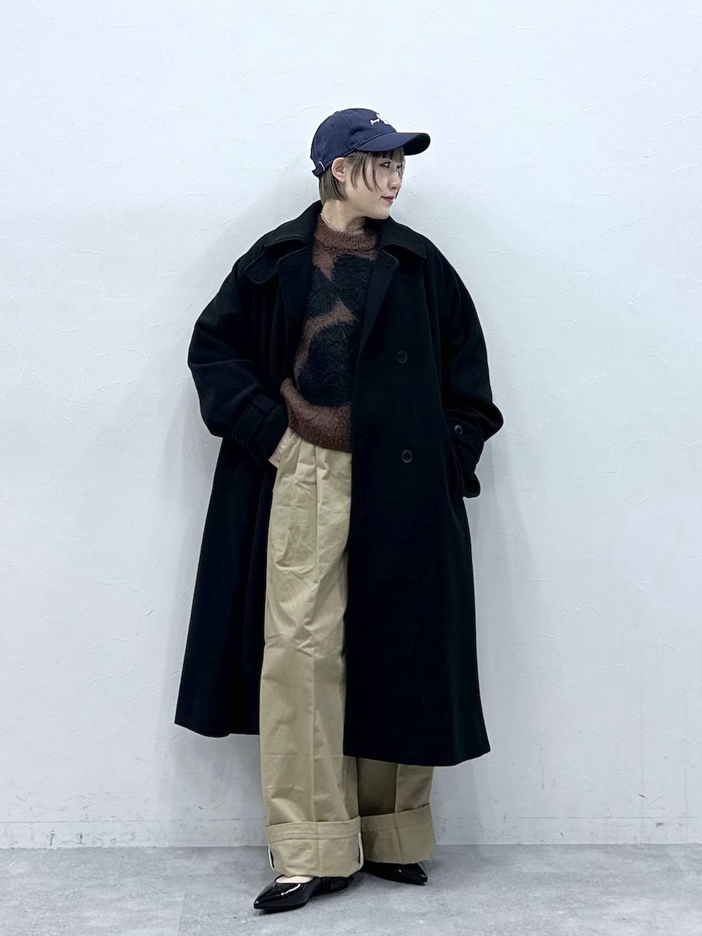 ブラック系のステンカラーコート、ブラウン系のニット/セーター、ベージュ系のチノパンツを着用したレディースの冬コーディネートの2枚目の写真