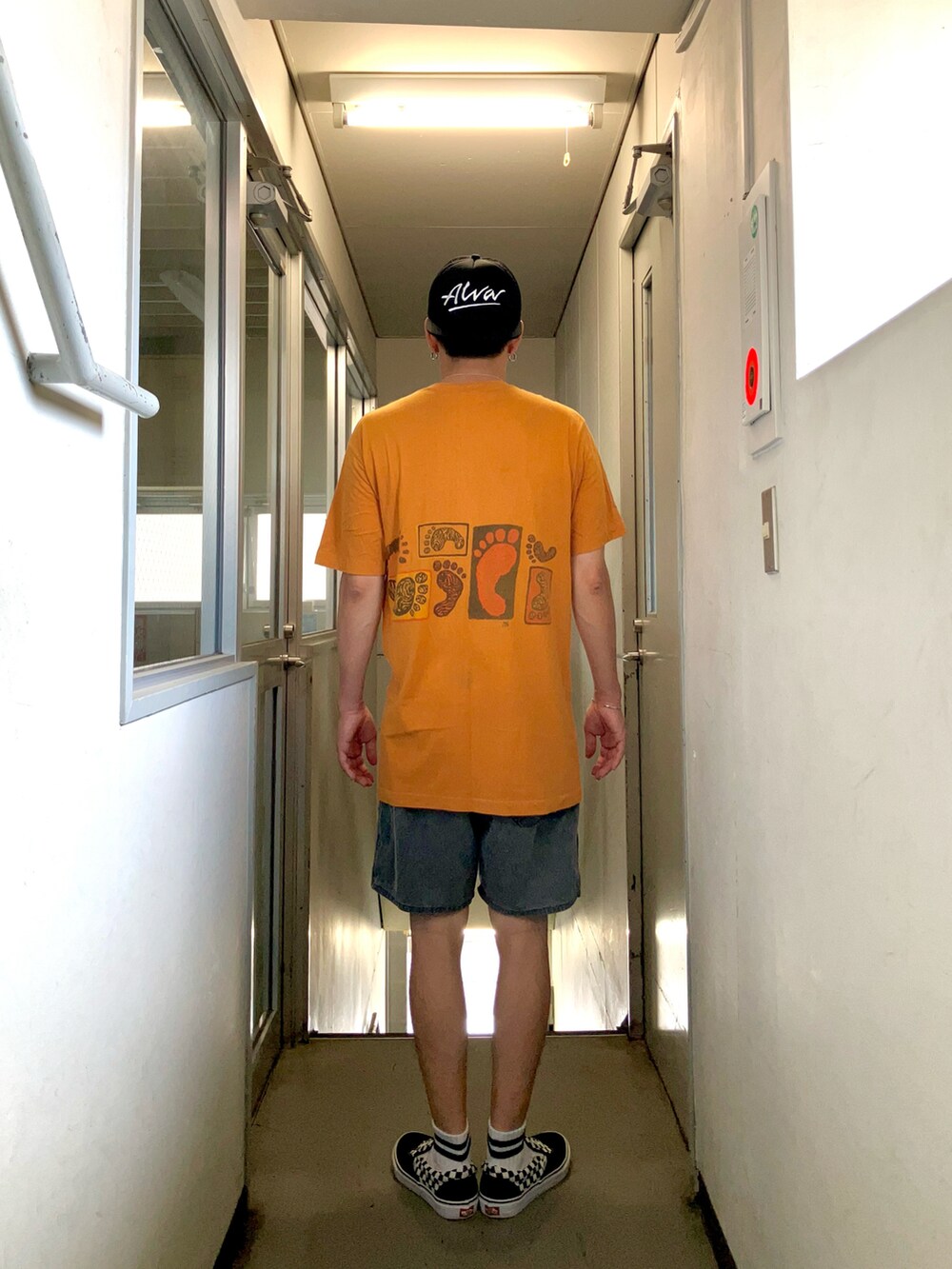 オレンジ系のTシャツ/カットソー、グレー系のパンツ、ブラック系のスニーカーを着用したメンズの夏コーディネートの2枚目の写真