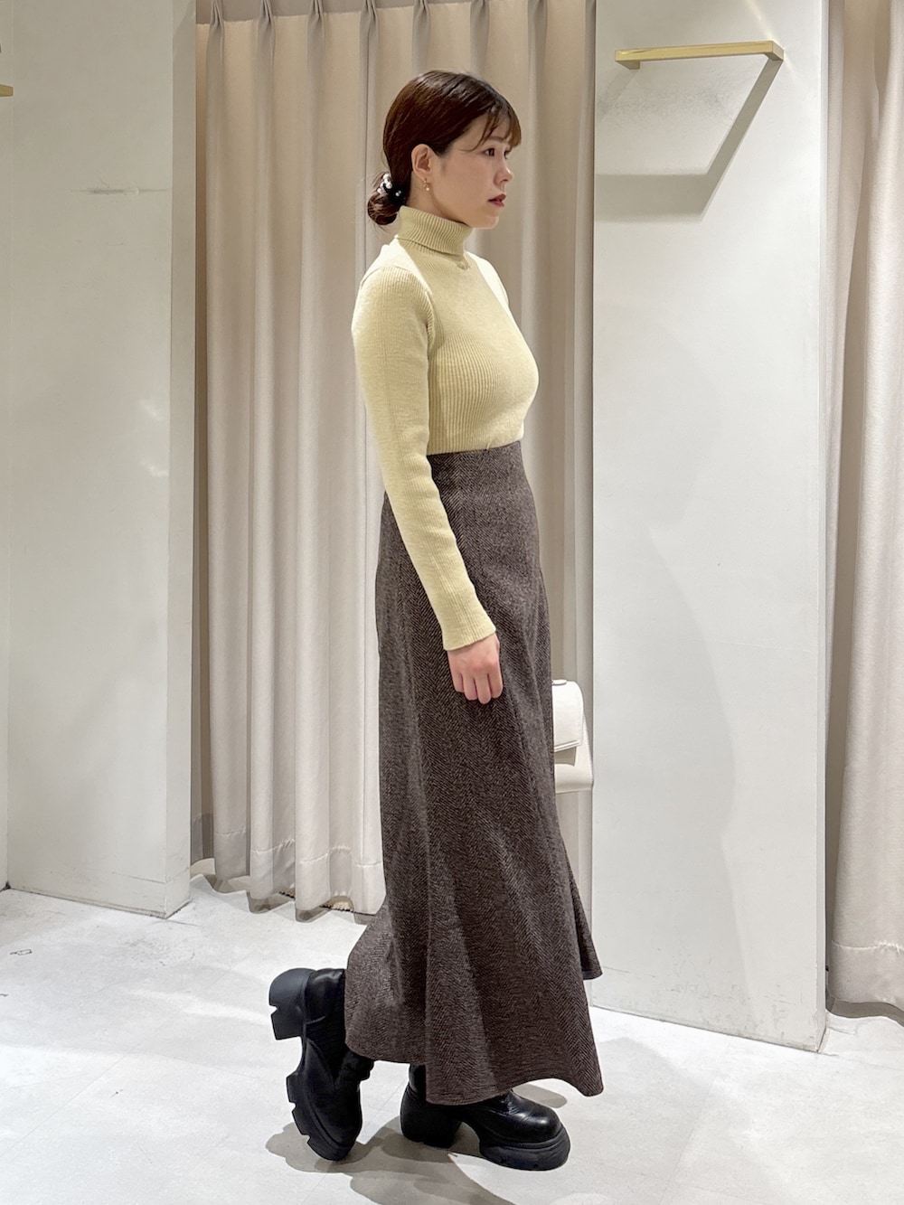 イエロー系のニット/セーター、ブラウン系のスカート、ブラック系のブーツを着用したレディースの秋コーディネートの2枚目の写真