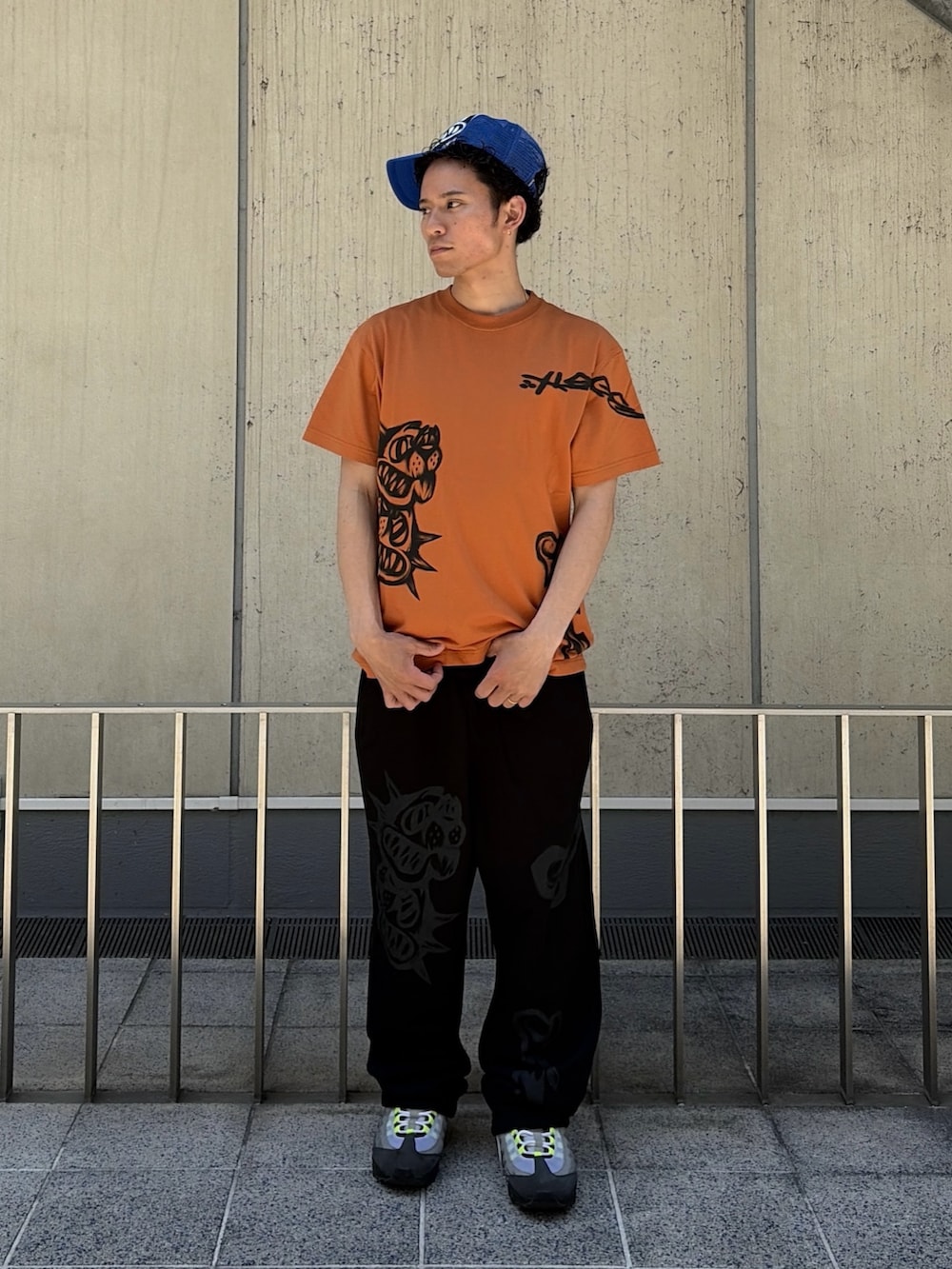オレンジ系のTシャツ/カットソー、ブラック系のスウェットパンツ、ブルー系のキャップを着用したメンズの春コーディネートの2枚目の写真