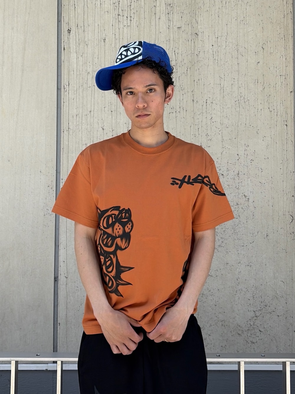 オレンジ系のTシャツ/カットソー、ブラック系のスウェットパンツ、ブルー系のキャップを着用したメンズの春コーディネートの6枚目の写真