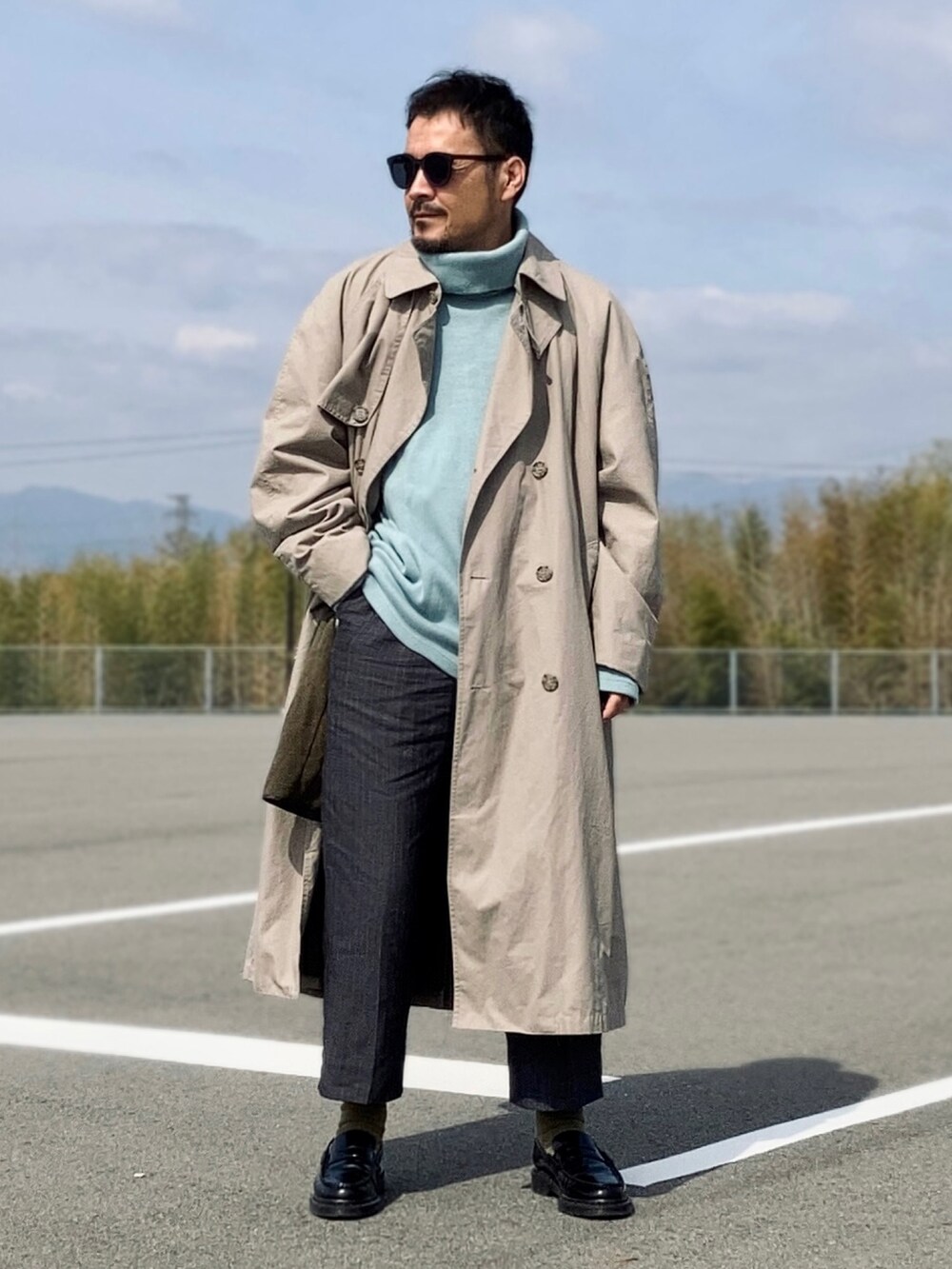 ブルー系のニット/セーター、ベージュ系のトレンチコート、グレー系のスラックスを着用したメンズの春コーディネートの2枚目の写真