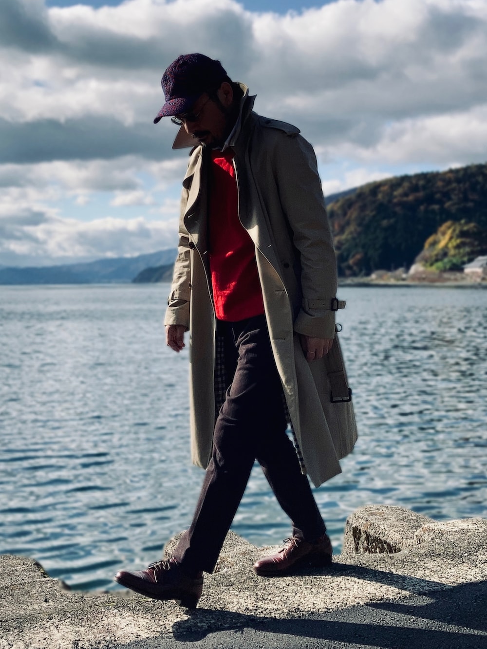 レッド系のニット/セーター、ホワイト系のシャツ/ブラウス、ベージュ系のトレンチコートを着用したメンズの秋コーディネートの2枚目の写真