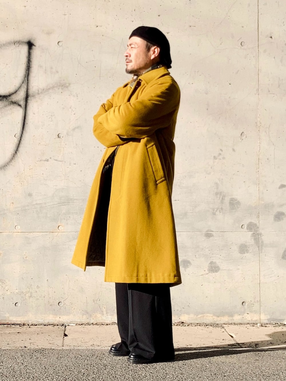 イエロー系のステンカラーコート、ブラック系のスラックス、ブラック系のローファーを着用したメンズの冬コーディネートの2枚目の写真