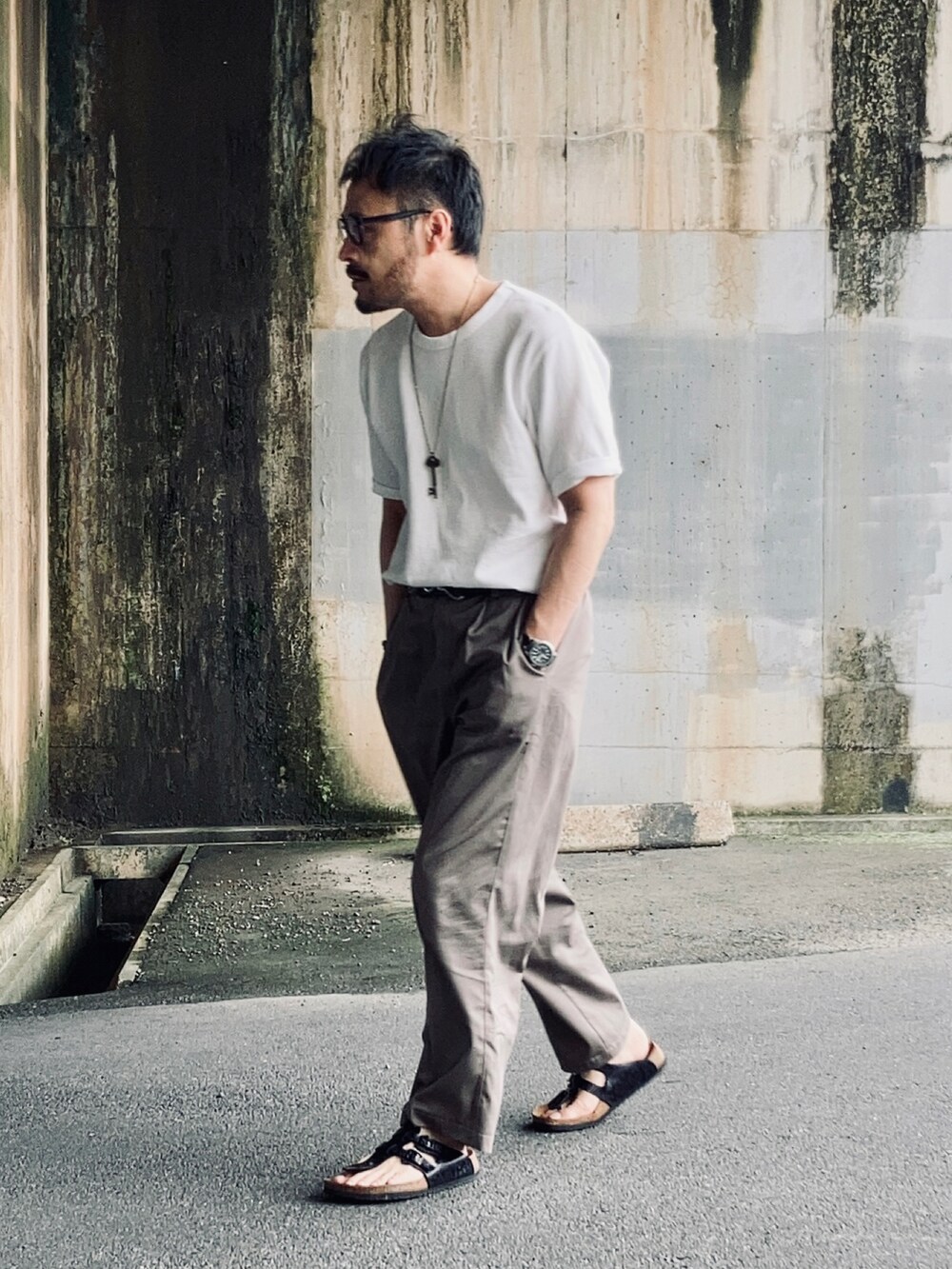 ホワイト系のTシャツ/カットソー、グレー系のスラックス、ブラック系のサンダルを着用したメンズの夏コーディネートの3枚目の写真