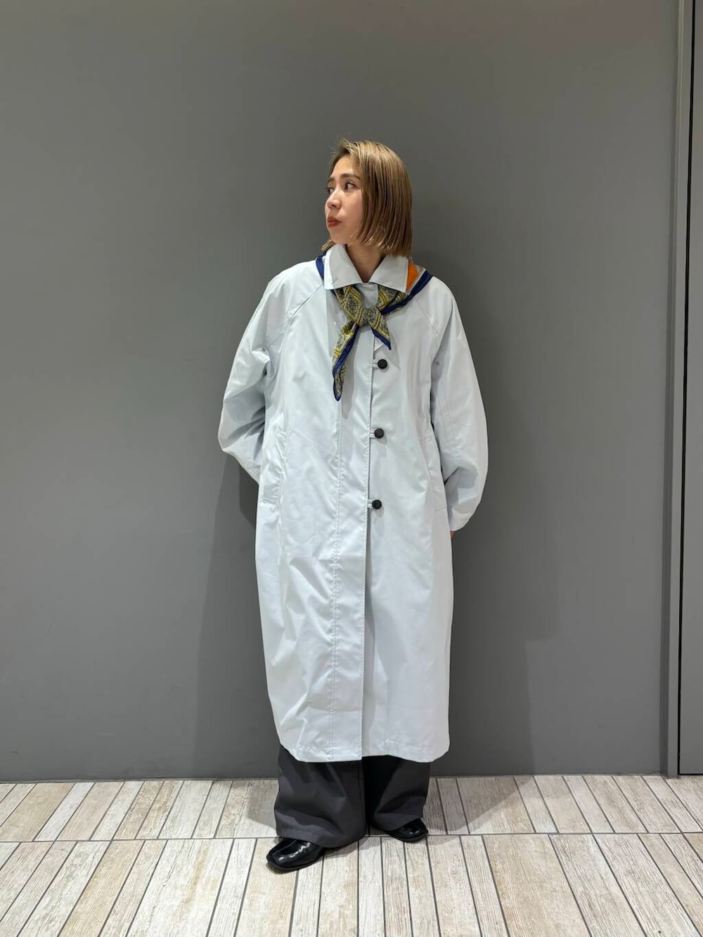 グレー系のスラックス、ブルー系のステンカラーコート、イエロー系のバンダナ/スカーフを着用したレディースの冬コーディネートの2枚目の写真