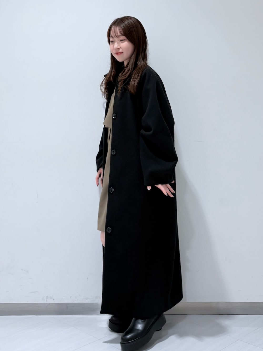 ブラック系のステンカラーコート、ブラウン系のワンピース、ブラック系のブーツを着用したレディースの秋コーディネートの2枚目の写真