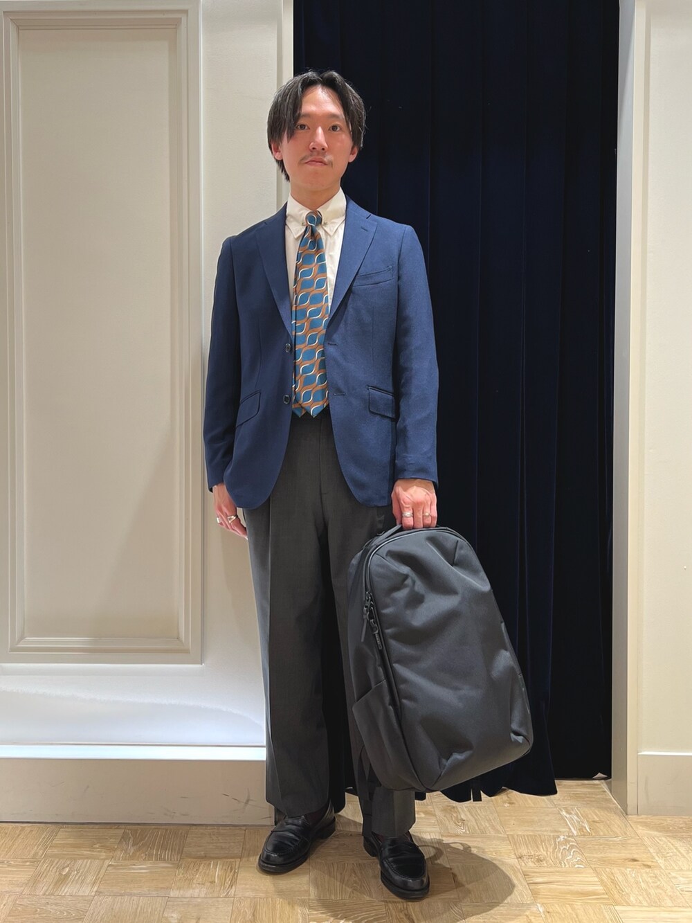 ブルー系のネクタイ、ブルー系のテーラードジャケット、ブラック系のバックパック/リュックを着用したメンズの春コーディネートの2枚目の写真
