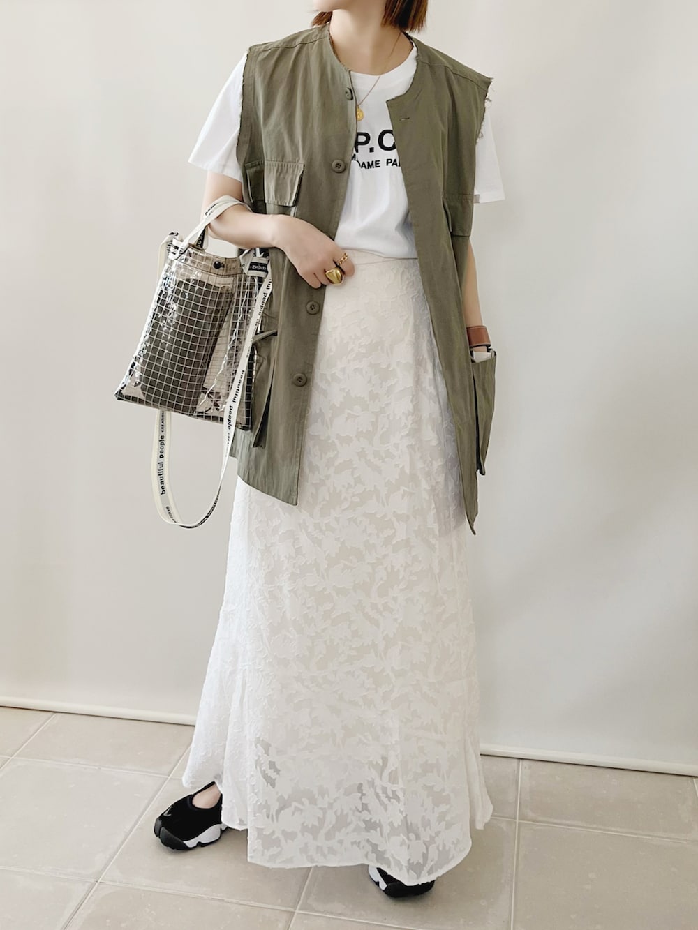グリーン系のベスト、ホワイト系のスカート、ブラック系のショルダーバッグを着用したレディースの夏コーディネートの2枚目の写真