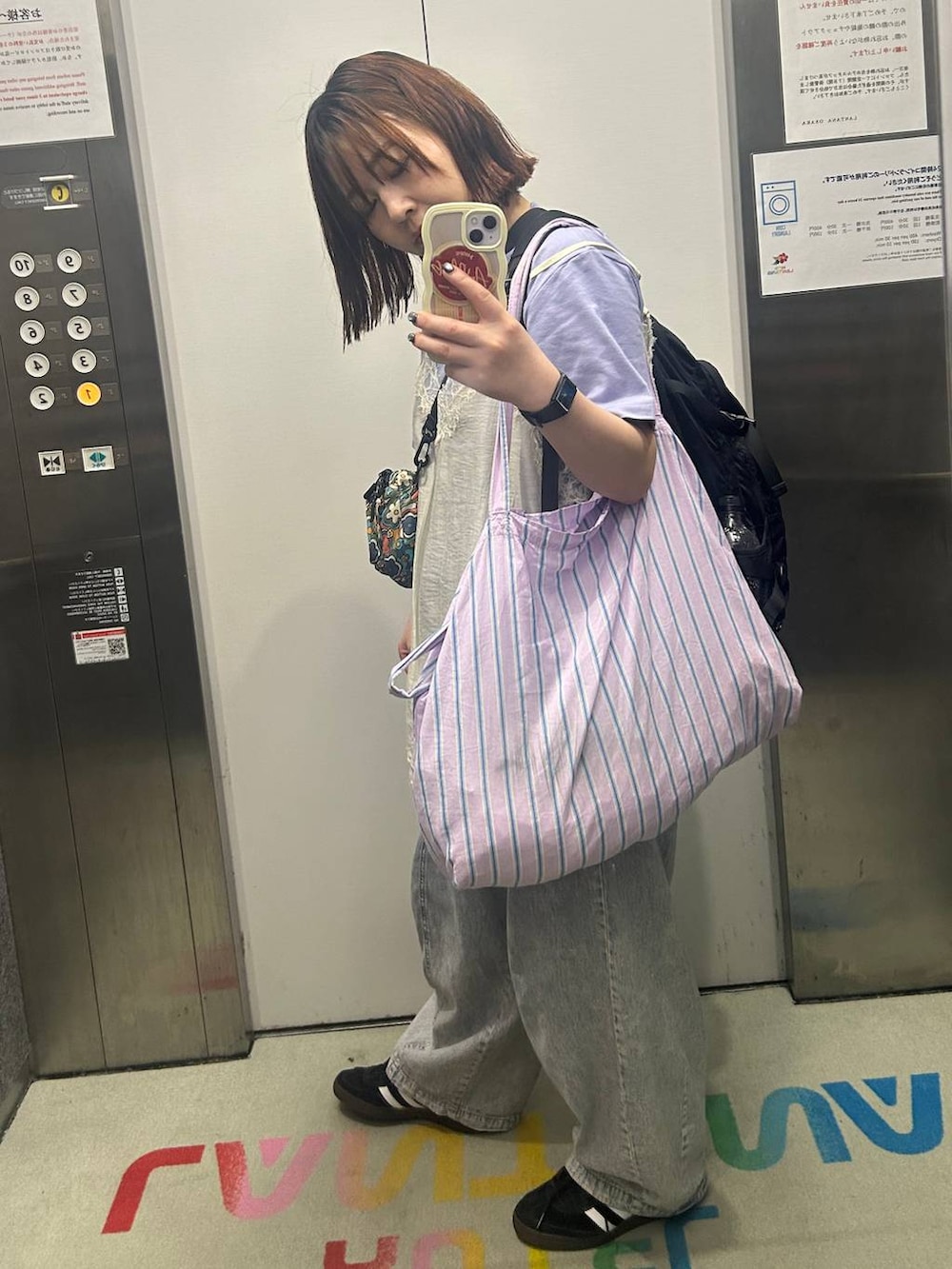 パープル系のトートバッグ、ブラック系のスニーカー、ブルー系のデニムパンツを着用したレディースの夏コーディネートの3枚目の写真