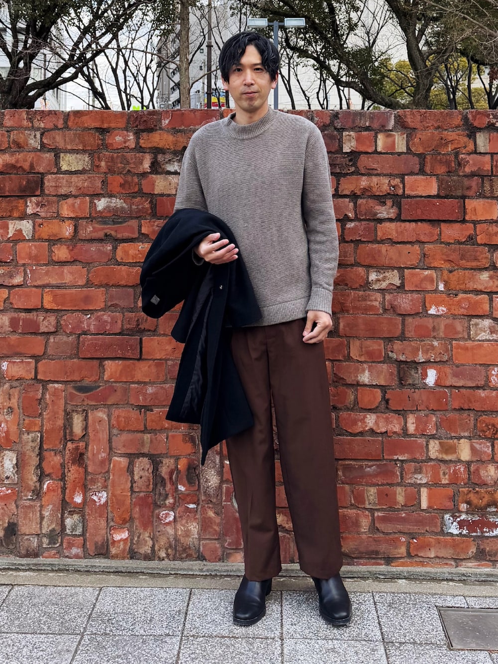 ブラック系のチェスターコート、グレー系のニット/セーター、ブラウン系のスラックスを着用したメンズの冬コーディネートの3枚目の写真