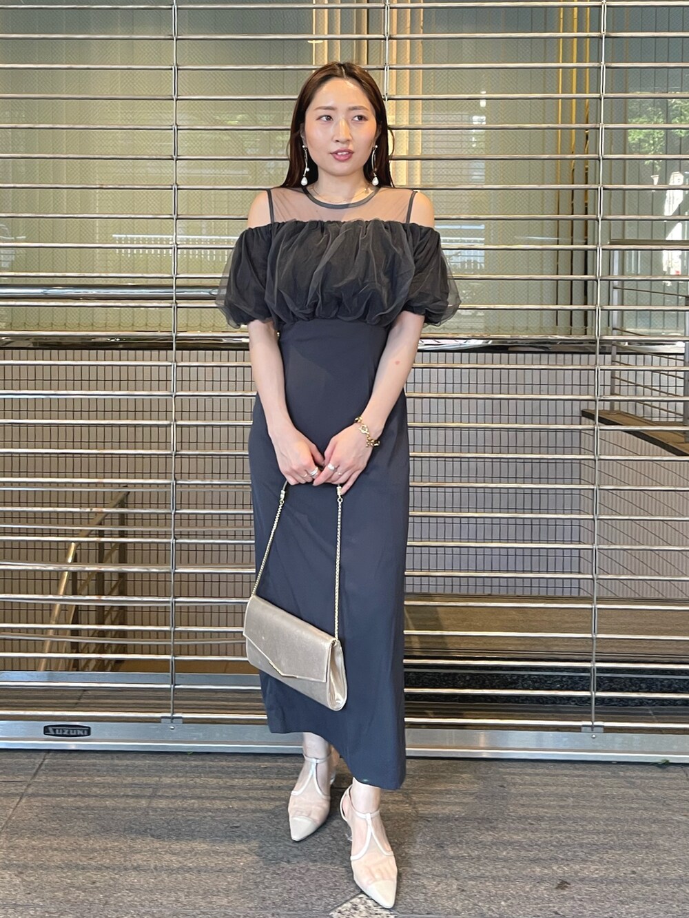 グレー系のドレス、ゴールド系のクラッチバッグ、ゴールド系のピアス（両耳用）を着用したレディースの夏コーディネートの2枚目の写真